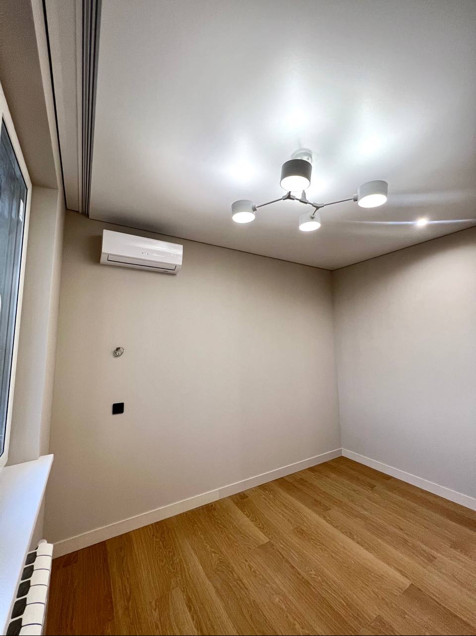 Pomalowane na beżowo puste wnętrze z drewnianą podłogą, nowoczesnym oświetleniem sufitowym i zamontowanym klimatyzatorem.