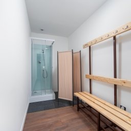 UCZNIOWSKI KLUB SPORTOWY ORLĘTA WROCŁAW - Wnętrze szatni z drewnianą ławką, wieszakami na ścianie, parawanem i kabiną prysznicową z chromowaną armaturą na tle białych ścian i brązowej podłogi.