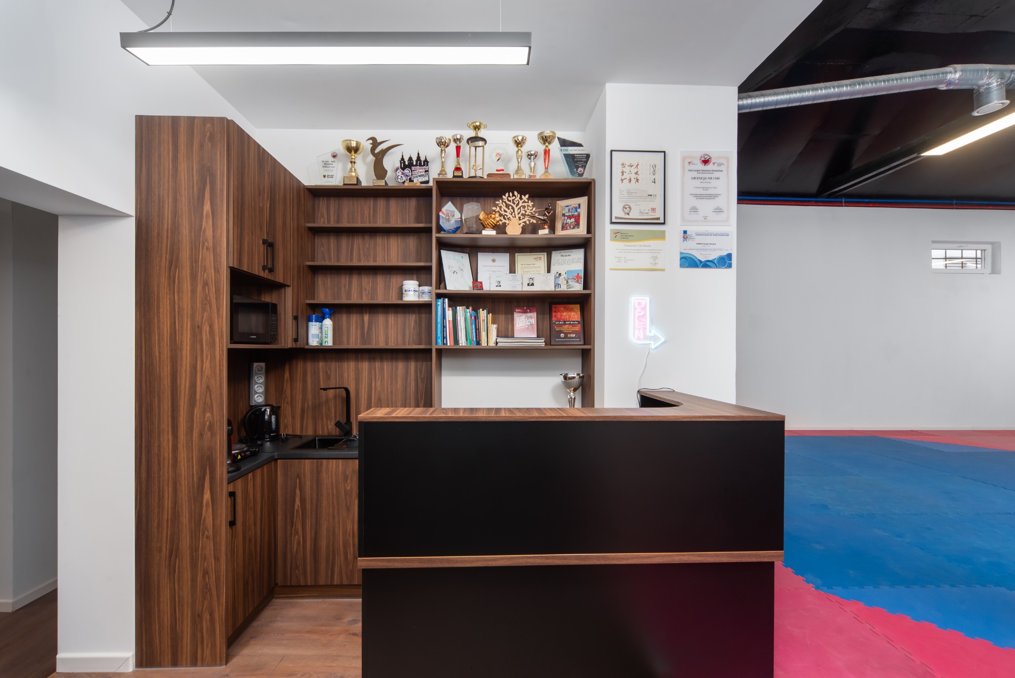 Recepcja w klubie sportów walki z trofeami na półkach, widoczna mata treningowa w tle.