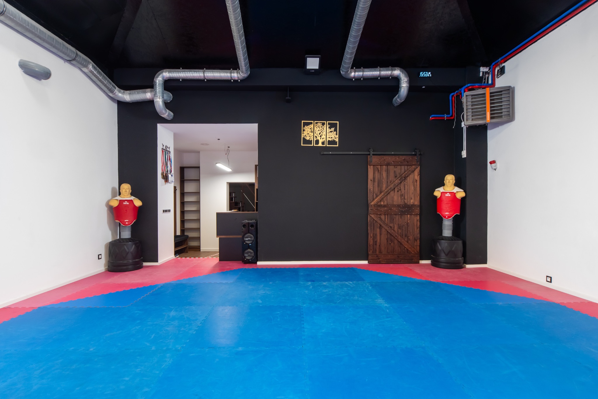 Sala treningowa z matą w kolorach niebieskim i czerwonym, manekiny treningowe, drewniane drzwi przesuwne i system wentylacji podsufitowej.