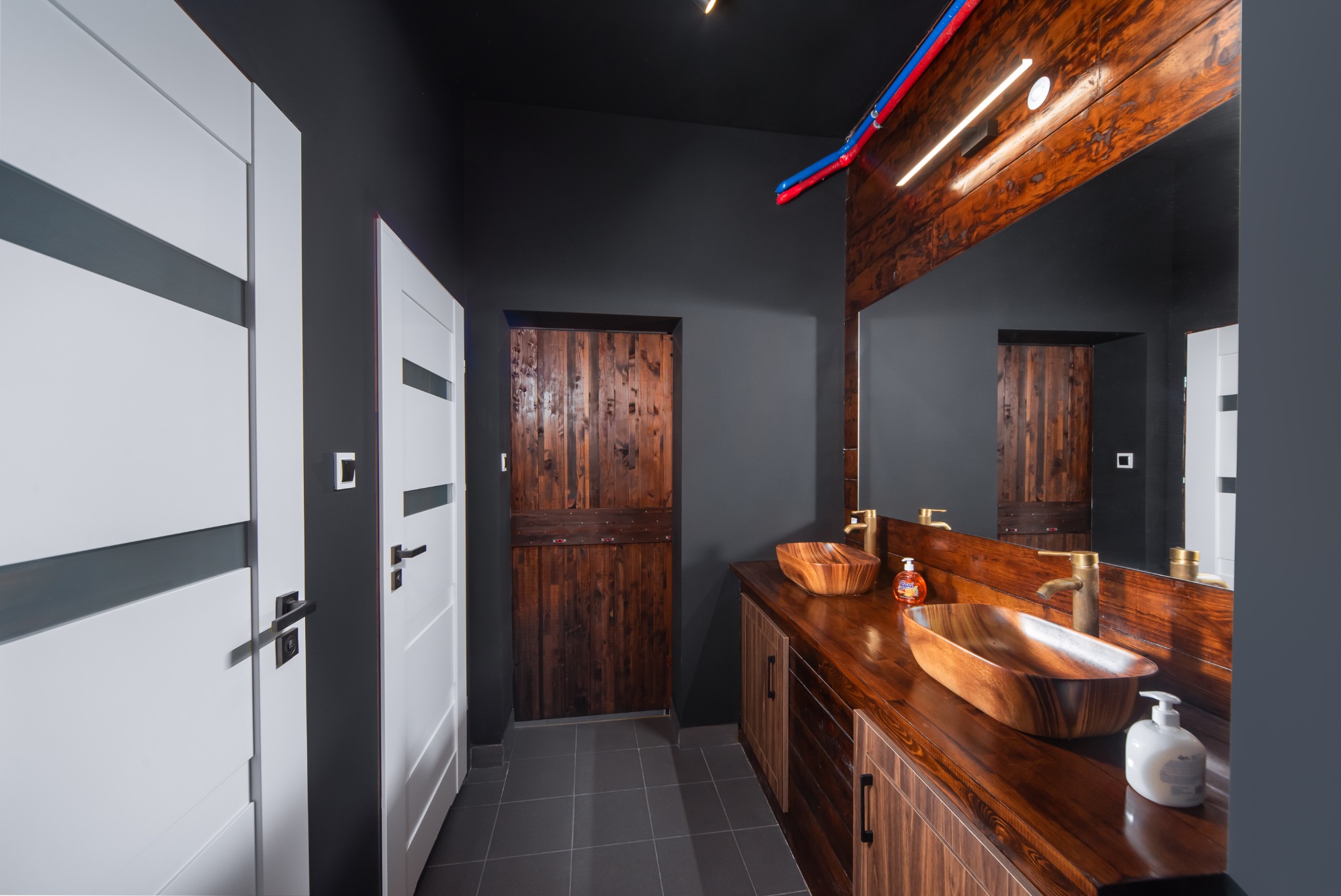 Nowoczesna łazienka z drewnianymi umywalkami w kształcie łodzi, blatem z ciemnego drewna i szarymi ścianami; widoczne białe drzwi z minimalistycznym designem i drewniane drzwi wejściowe.