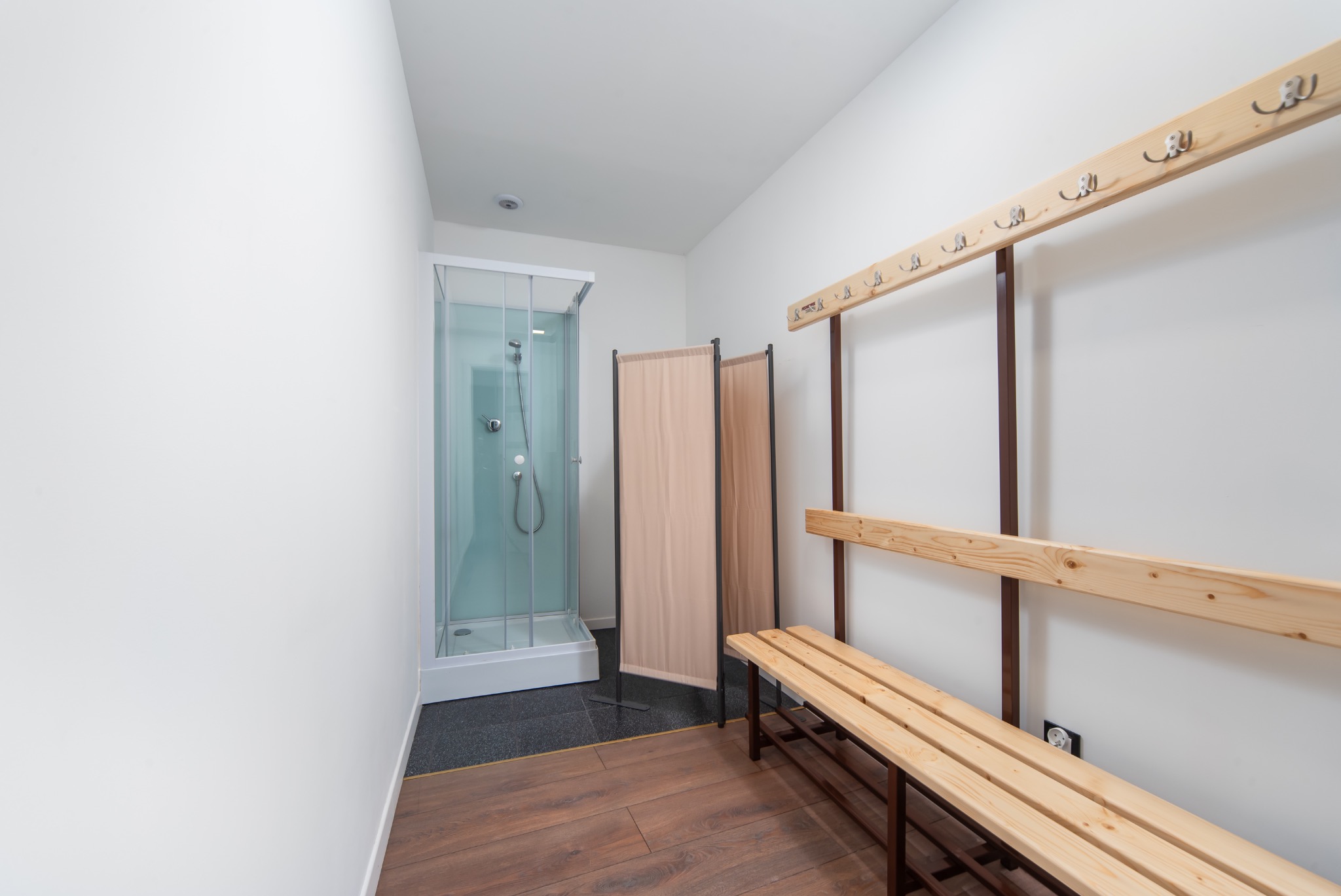 Wnętrze szatni z drewnianą ławką, wieszakami na ścianie, parawanem i kabiną prysznicową z chromowaną armaturą na tle białych ścian i brązowej podłogi.