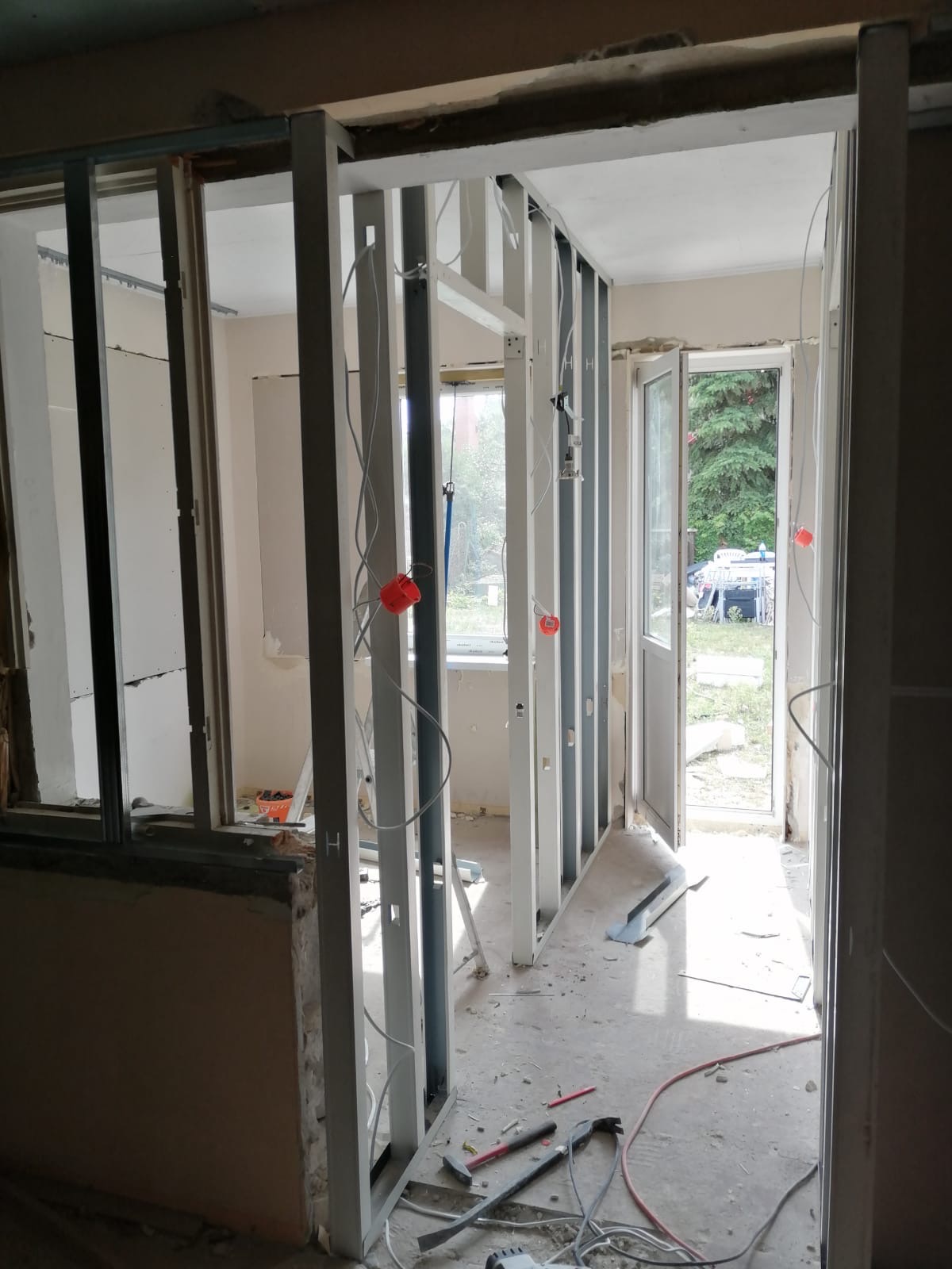 Wnętrze w trakcie tworzenia ścian działowych z metalowych profili, widoczne przewody elektryczne, narzędzia na podłodze i otwarte drzwi balkonowe z widokiem na zieleń.