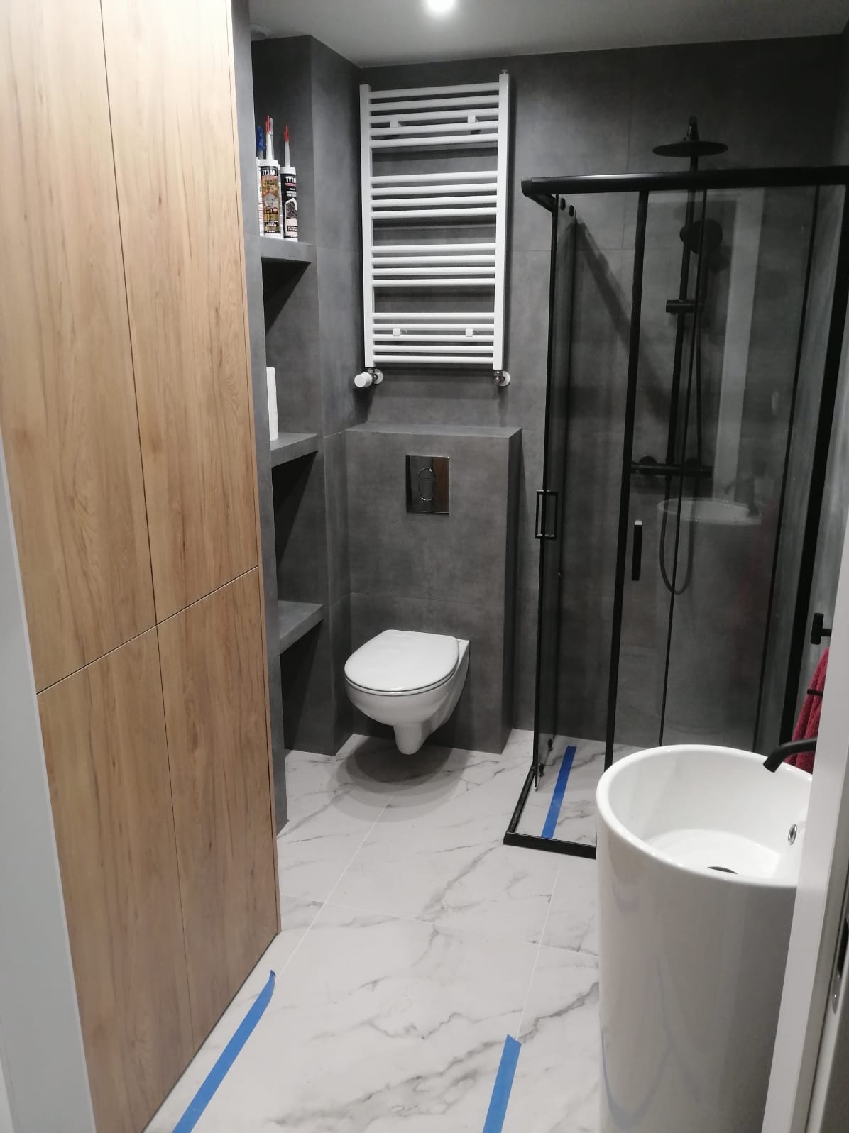 Nowoczesna łazienka z szarą glazurą, białą umywalką kolumnową, wiszącą toaletą i kabiną prysznicową z czarnymi ramami, zabezpieczona niebieską taśmą malarską.