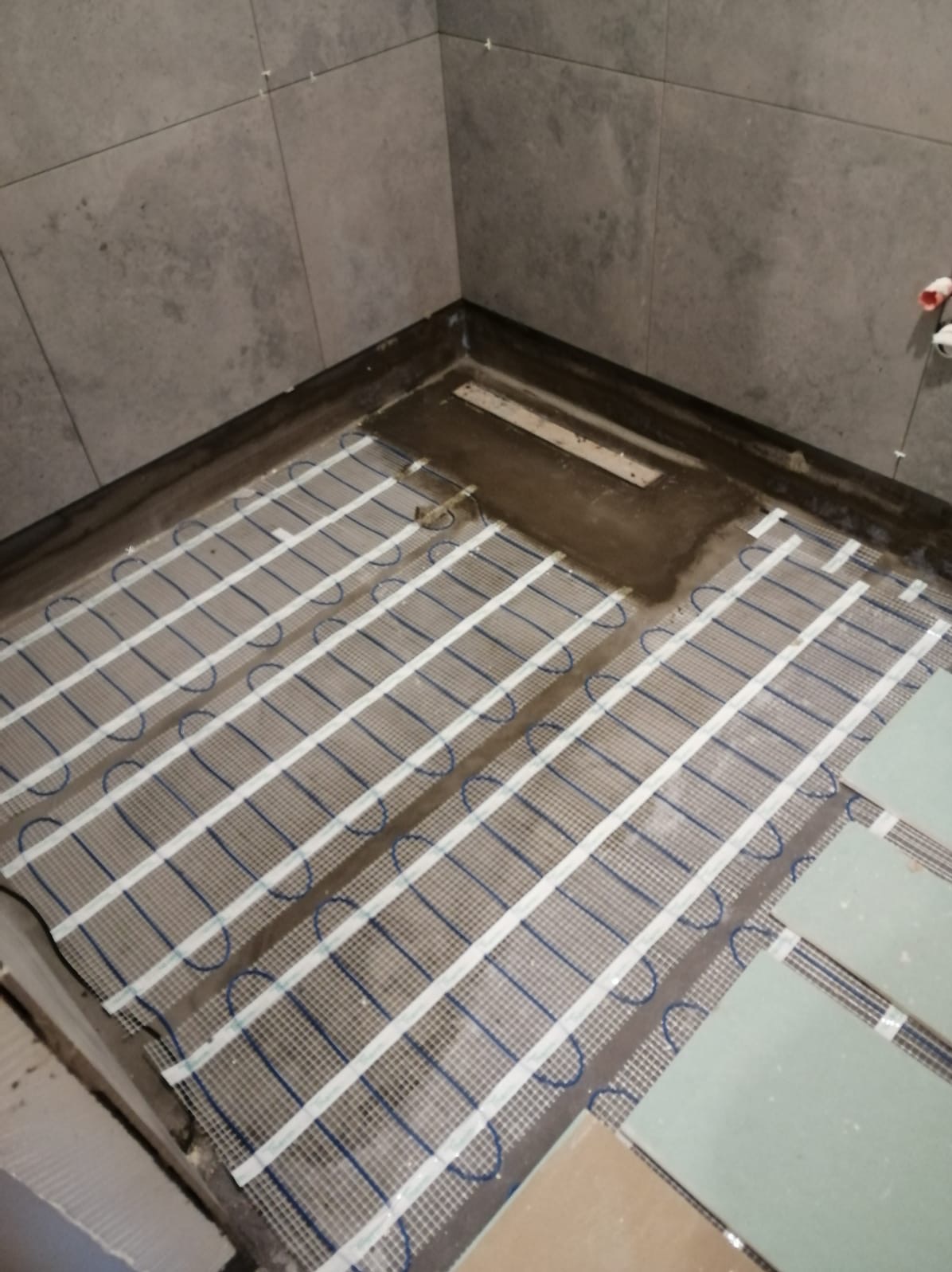 Montaż elektrycznego ogrzewania podłogowego na siatce w przygotowanej do wykończenia łazience, widoczny odpływ liniowy.