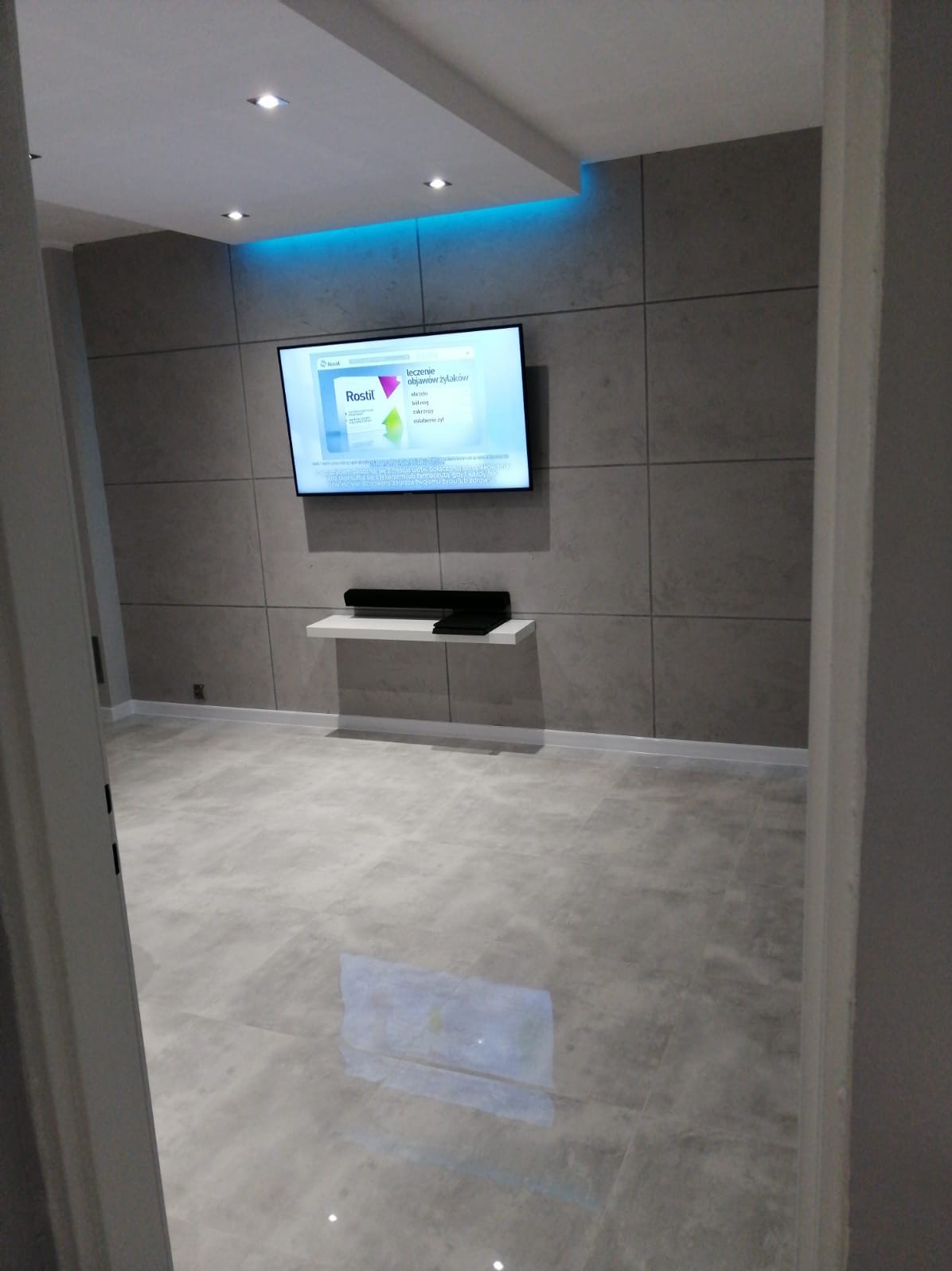 Salon z szarymi płytkami na ścianach i podłodze, podświetleniem LED na suficie i telewizorem na ścianie.