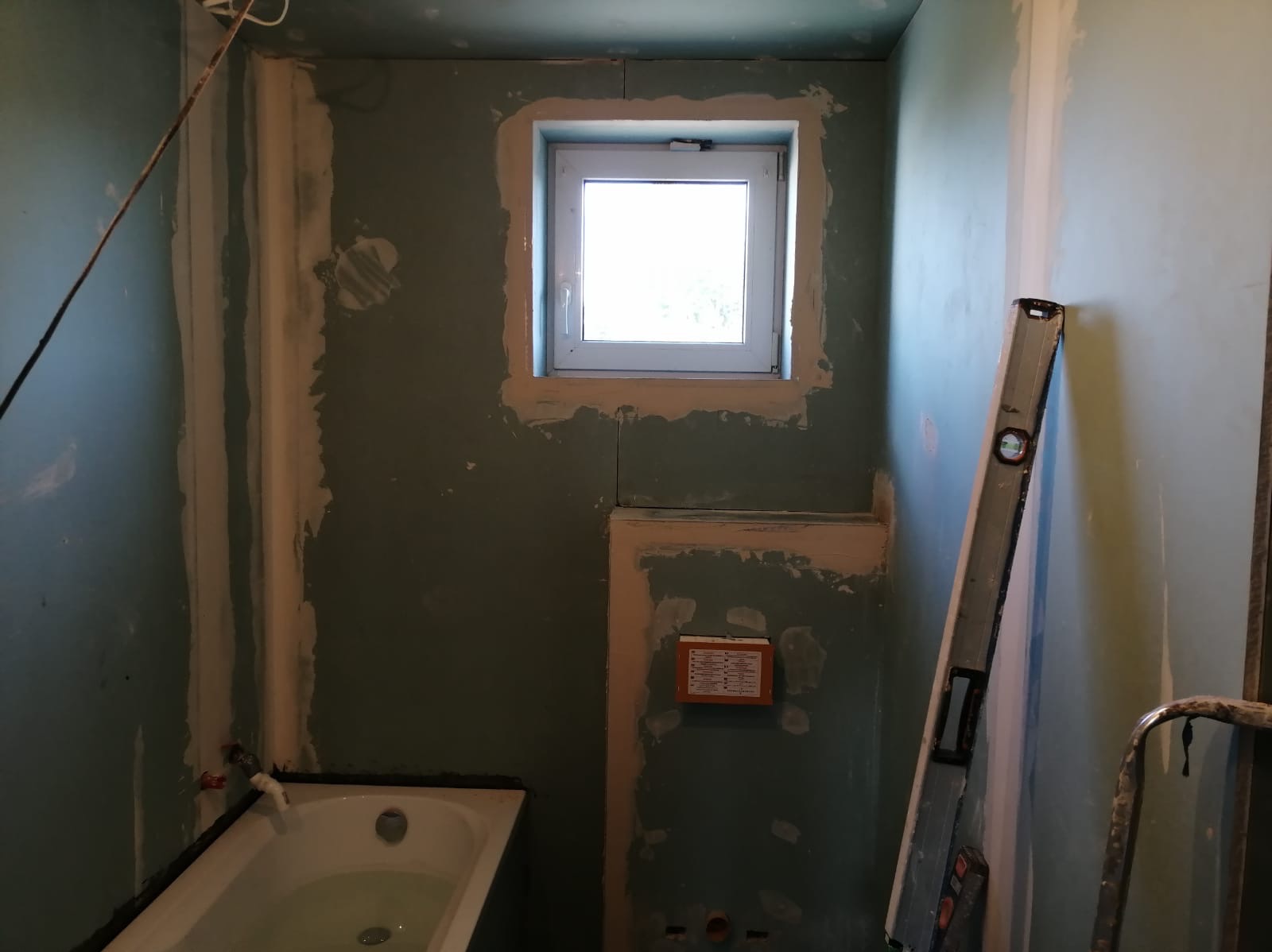 Przygotowanie ścian podłoża w łazience: płyty gipsowo-kartonowe, szpachlowanie przy oknie i wzdłuż rur, poziomica oparta o ścianę, częściowo widoczna wanna.