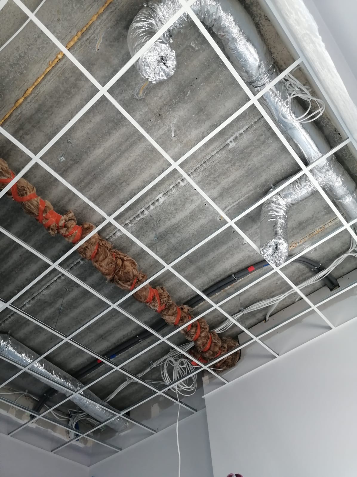Sufit podwieszany w trakcie montażu z widocznymi przewodami wentylacyjnymi, rurami i kablami, częściowo zaizolowanymi, zamocowanymi do sufitu konstrukcyjnego.