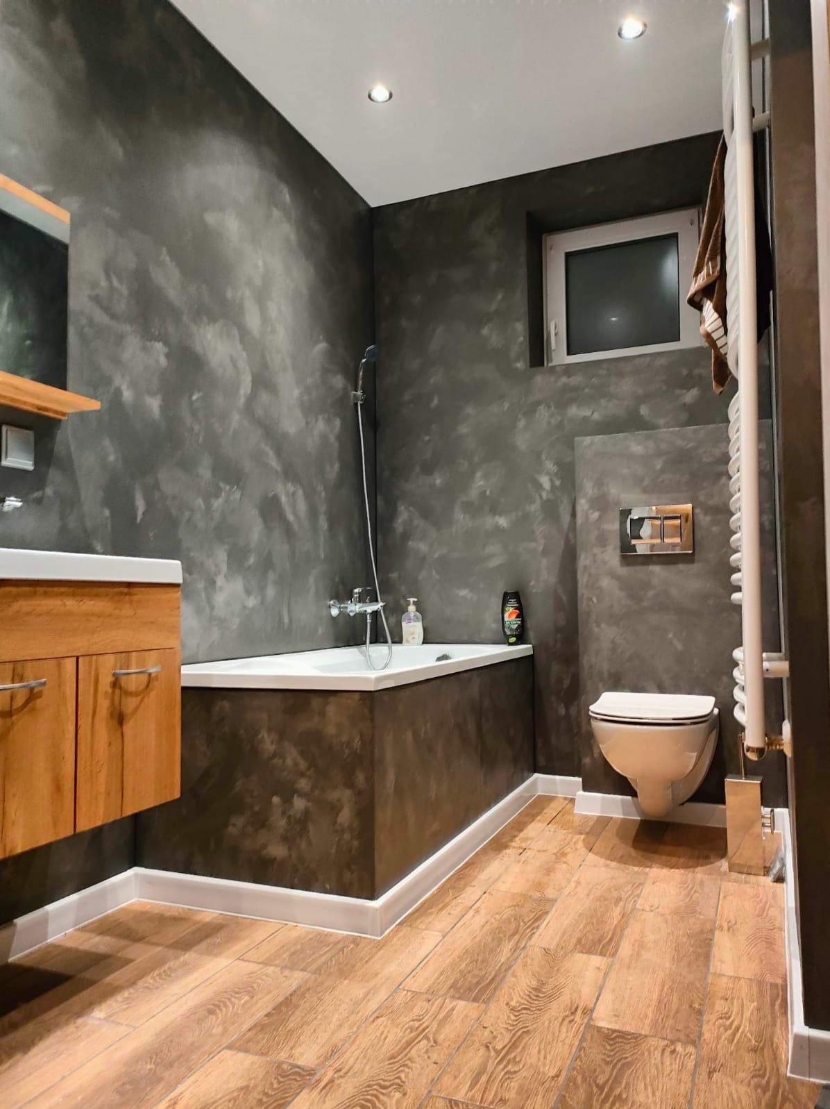 Nowoczesna łazienka z drewnianą podłogą, ścianami w odcieniach szarości z efektem dekoracyjnym, białą wanną obudowaną w tonacji ścian i wiszącą toaletą.