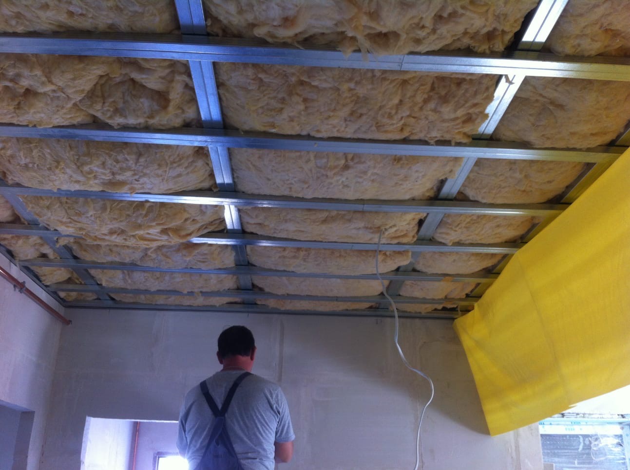 Montaż izolacji z wełny mineralnej w metalowej konstrukcji sufitu podwieszanego, widoczny pracownik w kombinezonie.