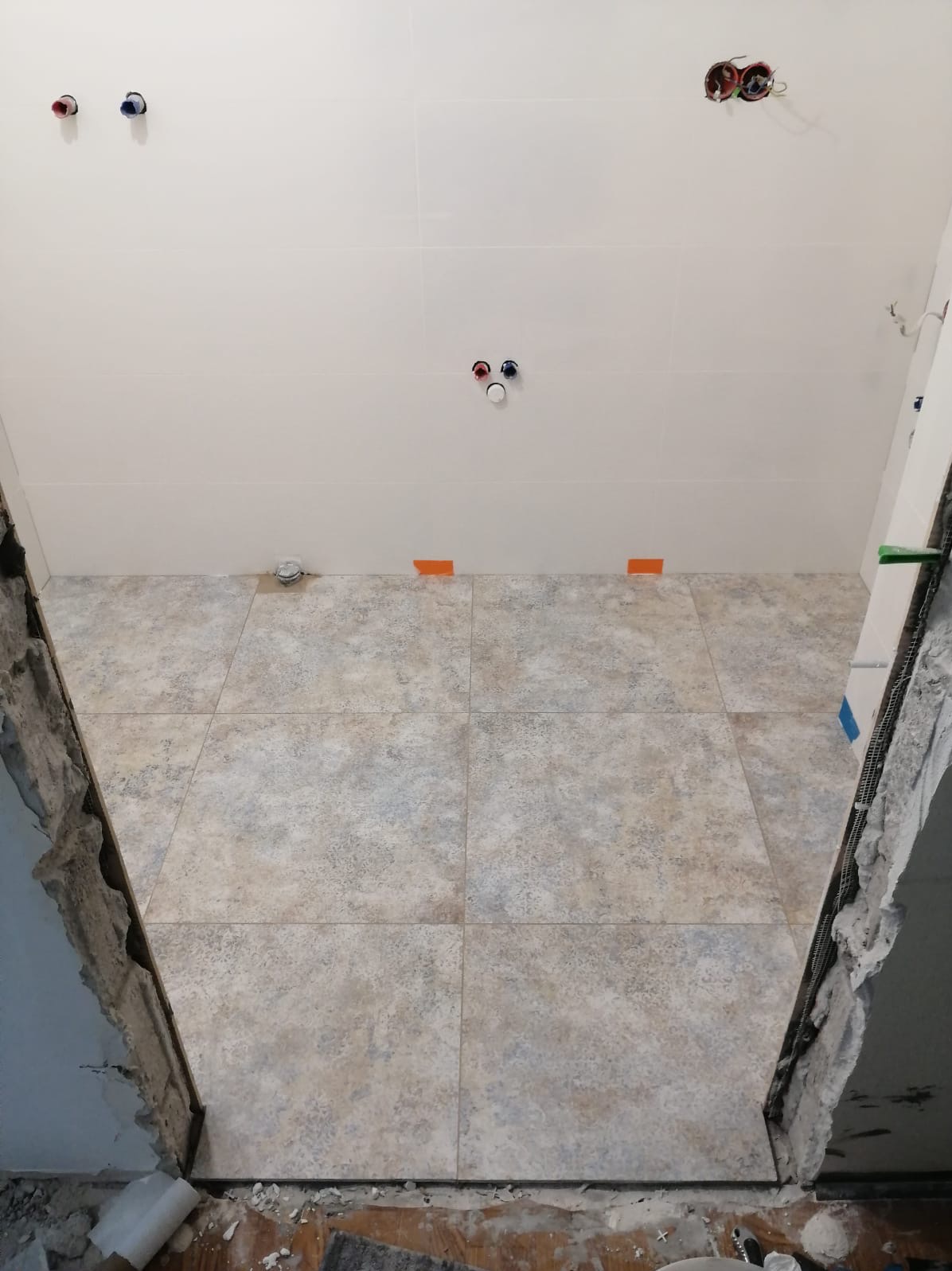 Łazienka w trakcie remontu, widoczne nowe płytki podłogowe w odcieniach beżu i szarości, surowe ściany przygotowane do dalszych prac, wystające instalacje wodne i elektryczne.