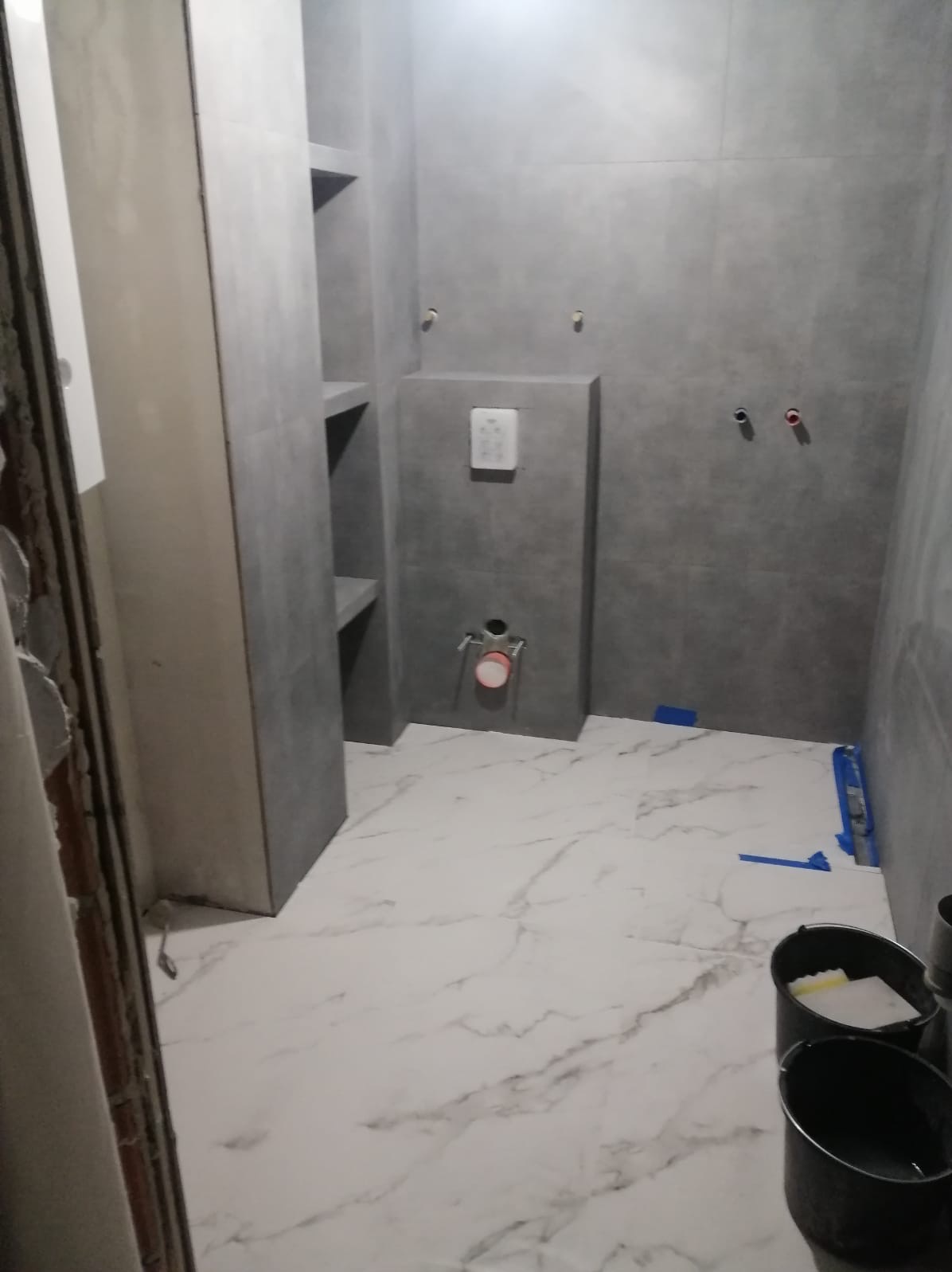 Wnętrze łazienki w trakcie prac wykończeniowych, widoczne szare płytki na ścianach, białe płytki imitujące marmur na podłodze, zabudowa WC, półki wnękowe, przygotowane do użycia wiadra i narzędzia...