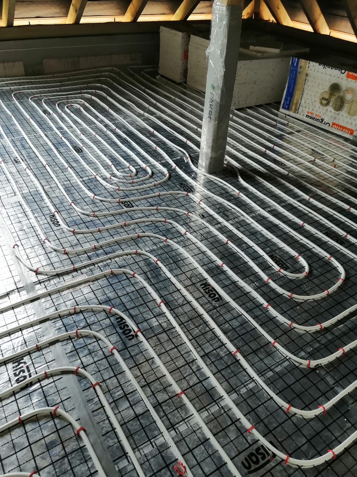 Białe rury ogrzewania podłogowego marki HISAN, ułożone na folii z czarną siatką, widoczne czerwone mocowania, w tle ocieplenie i słup owinięty folią.