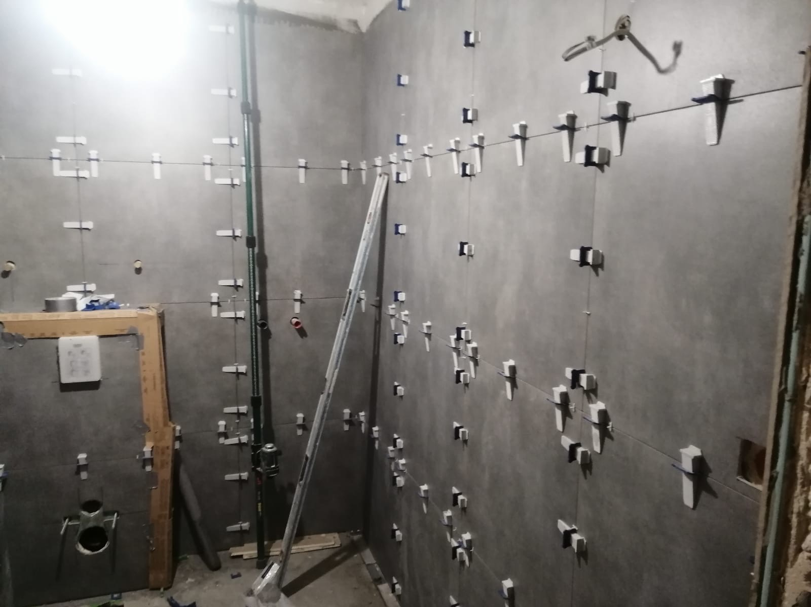 Ściana łazienki w trakcie układania szarych płytek wielkoformatowych, z użyciem systemu poziomowania płytek, widoczne pionowe rury instalacyjne i laserowy niwelator w rogu pomieszczenia.