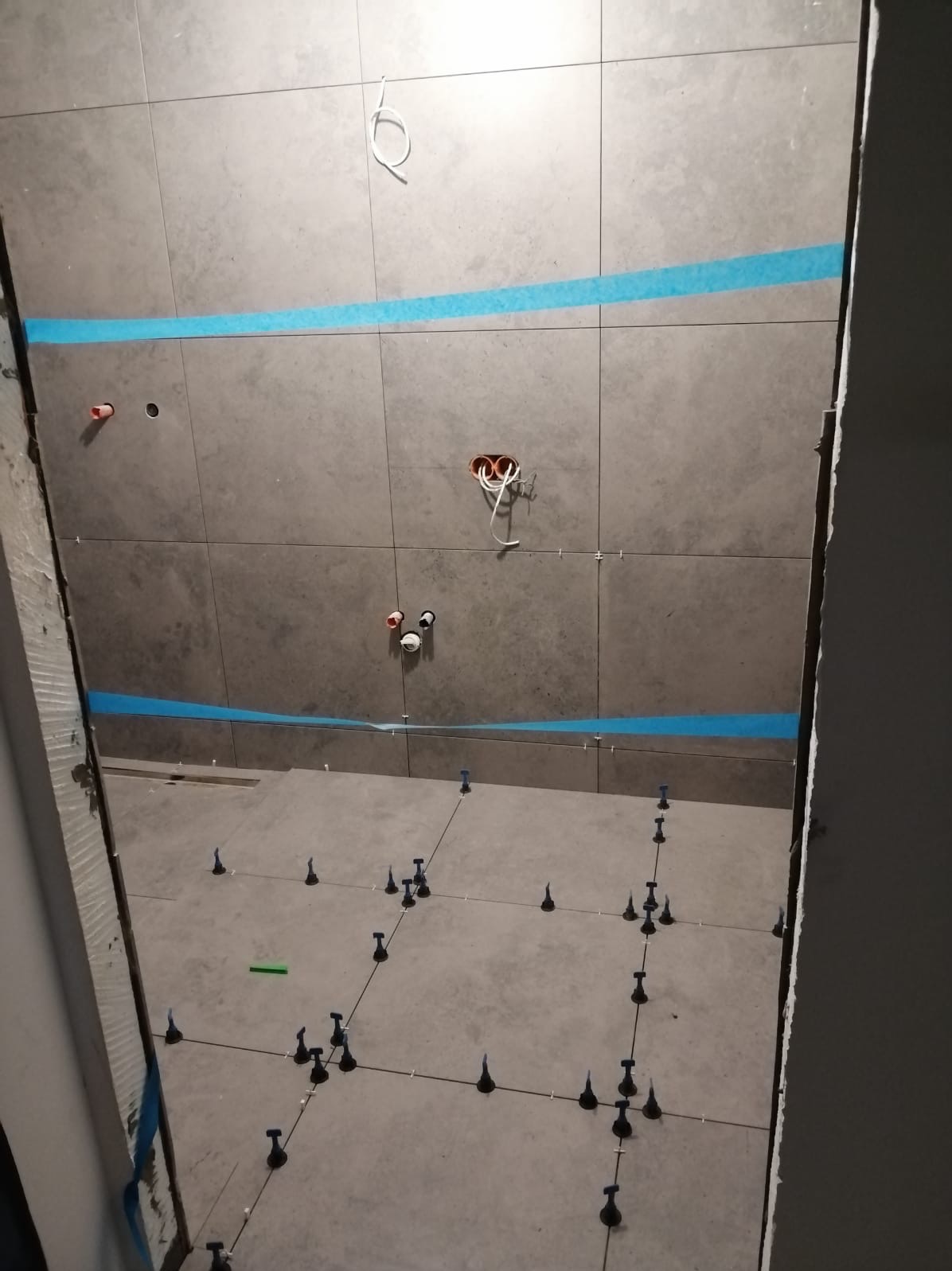 Układanie szarych płytek w łazience, widoczne niebieskie taśmy maskujące na ścianach, krzyżyki dystansowe i system poziomowania płytek na podłodze, przygotowane instalacje wodne i elektryczne.