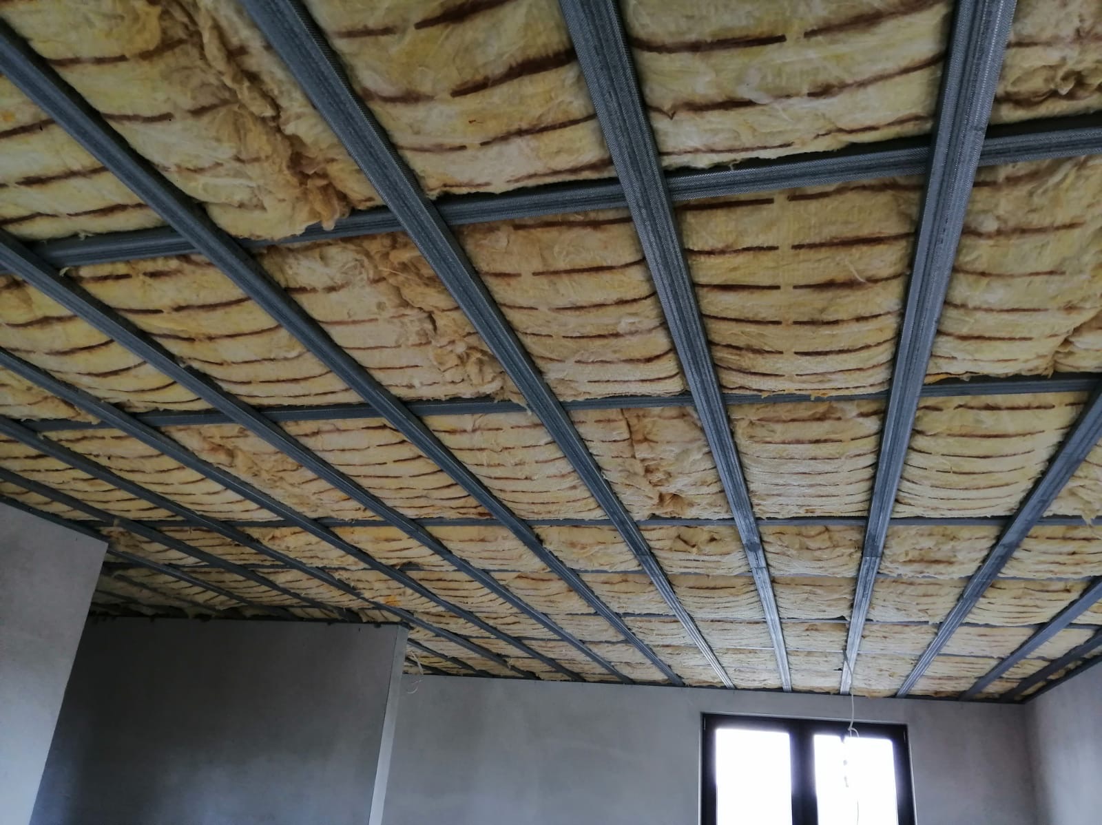 Sufit podwieszany w trakcie montażu, widoczne metalowe profile i warstwa izolacji z wełny mineralnej, surowy stan wykończenia wnętrza.