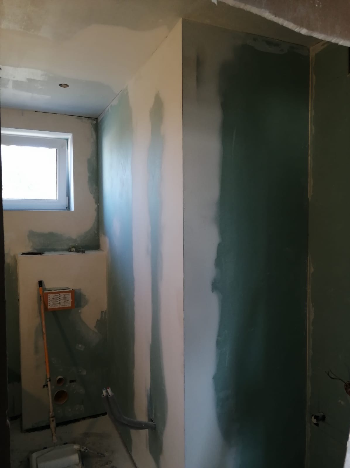 Wnętrze łazienki w trakcie prac wykończeniowych, widoczne ściany po szpachlowaniu, z fragmentami nałożonej gładzi, okno, instalacja sanitarna oraz narzędzia malarskie.