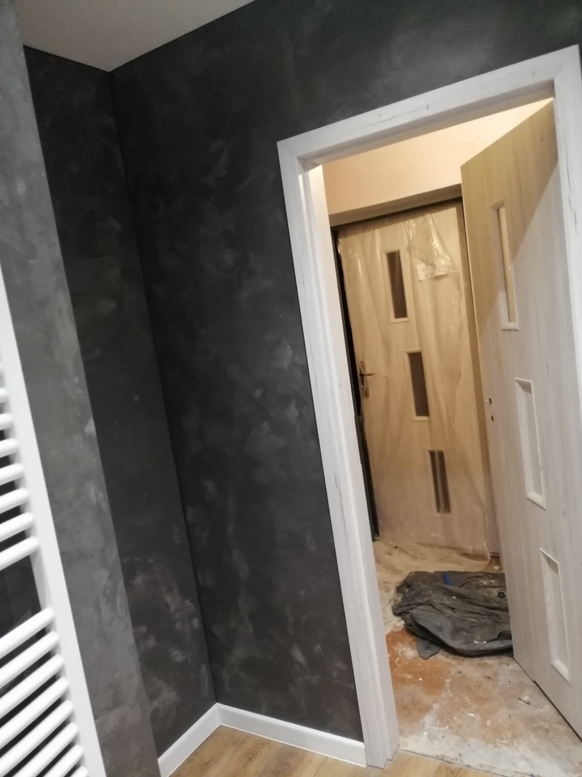 Ściana w ciemnoszarym odcieniu z efektem dekoracyjnym, widoczny biały grzejnik i fragment podłogi, w tle drzwi zabezpieczone folią ochronną.