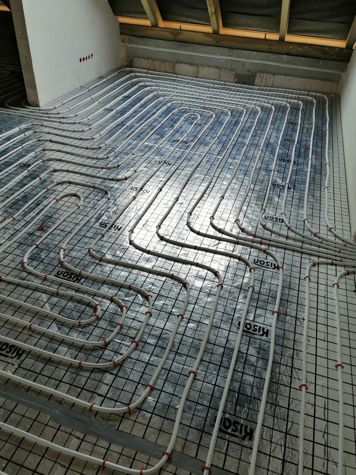 Białe rury ogrzewania podłogowego ułożone na srebrnej folii z nadrukiem kratki i logo producenta, widoczne elementy konstrukcji stropu.