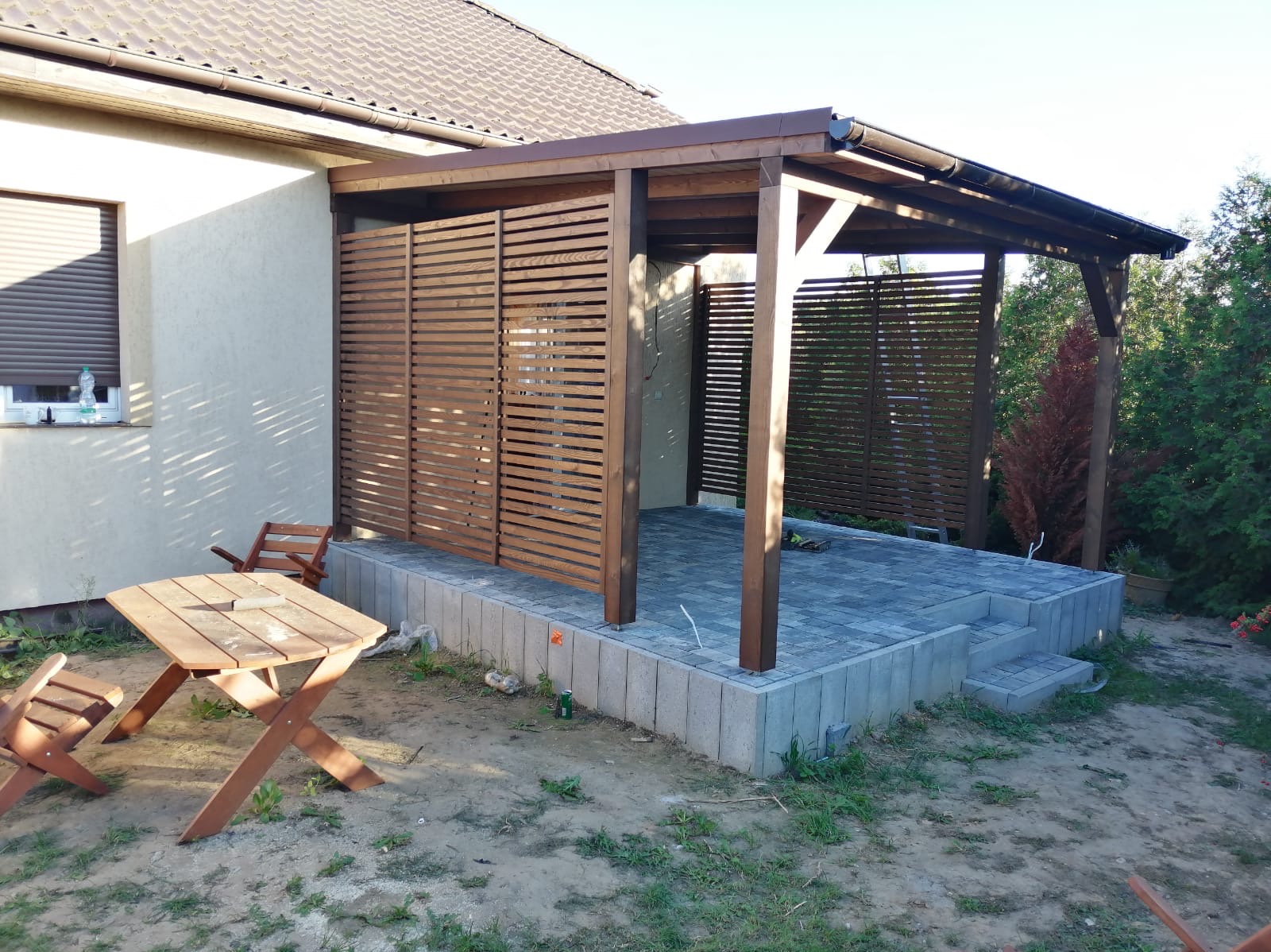 Drewniana altana z lamelami przylegająca do budynku, wyłożona kostką brukową, z drewnianym stołem i krzesłami na zewnątrz.