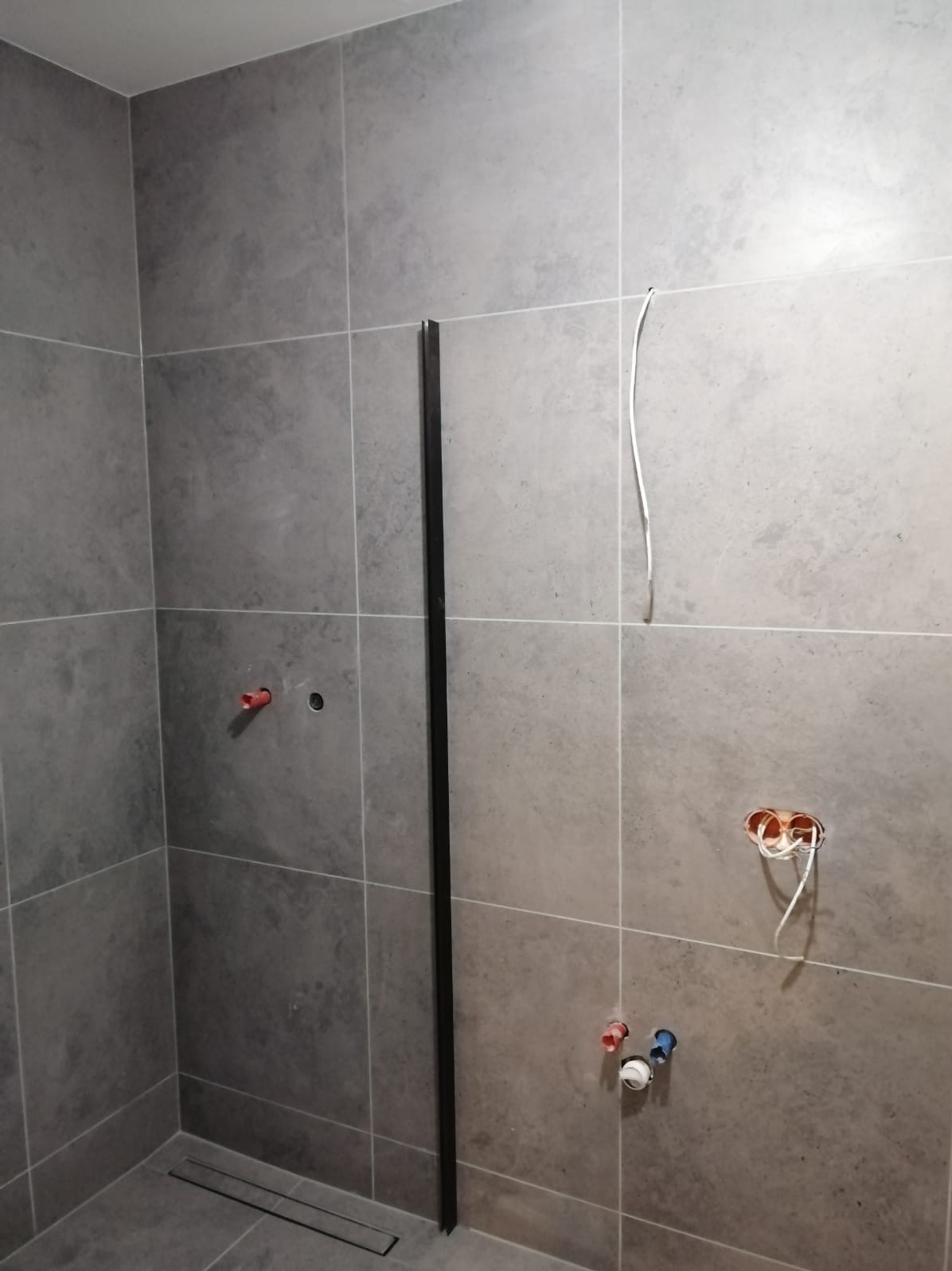 Szare płytki w trakcie montażu w łazience, widoczne wystające kable elektryczne i rury wodne, czarna listwa przyścienna.