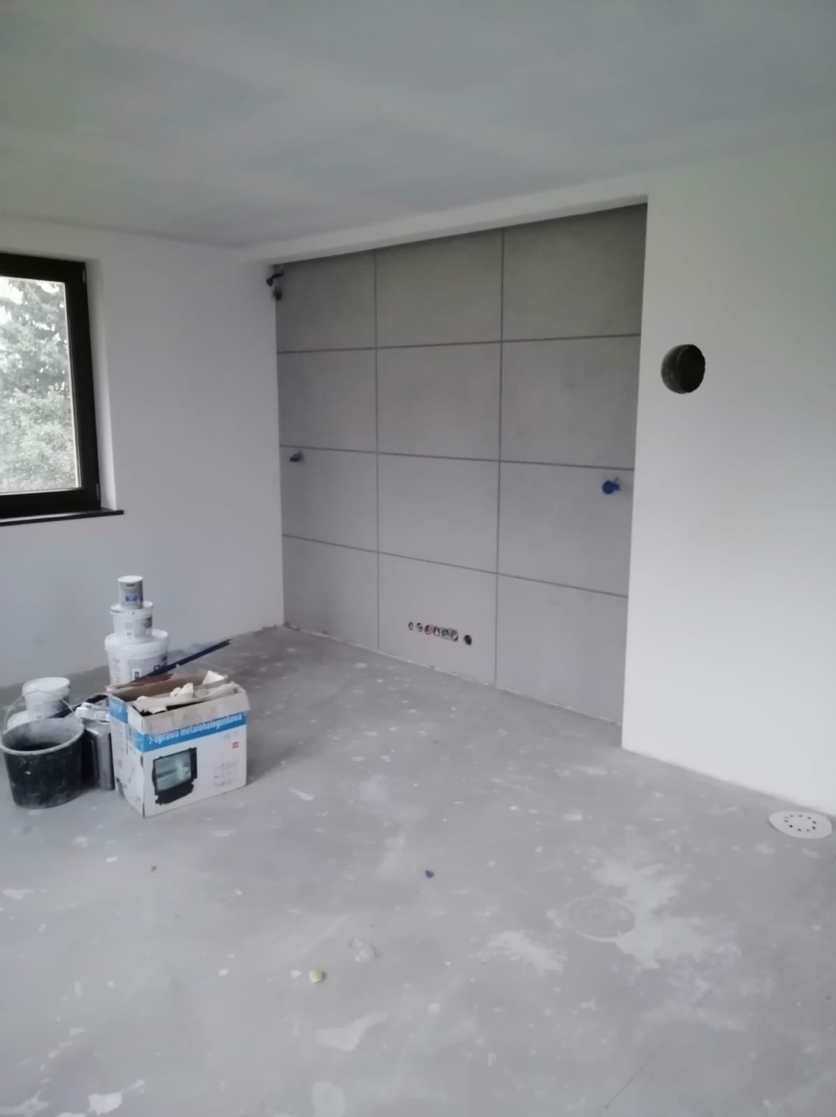 Łazienka w trakcie remontu: ściana z szarych płytek częściowo ułożona, widoczne otwory na instalacje, podłoga z resztkami zaprawy, puszki z farbą i narzędzia malarskie przy oknie.