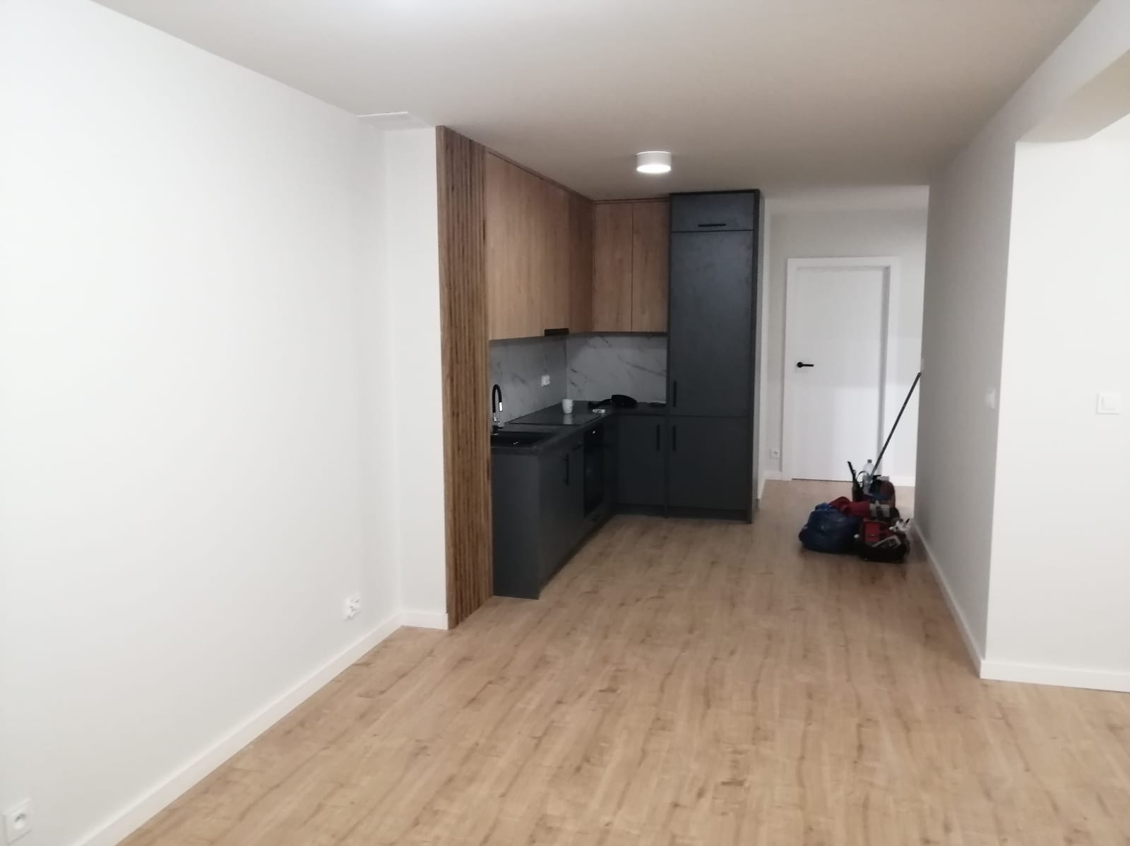 Nowoczesna, minimalistyczna kuchnia z drewnianymi i grafitowymi szafkami, marmurowym blatem i drewnianą ścianką działową, widok na białe ściany i podłogę z paneli, w tle sprzęt budowlany.