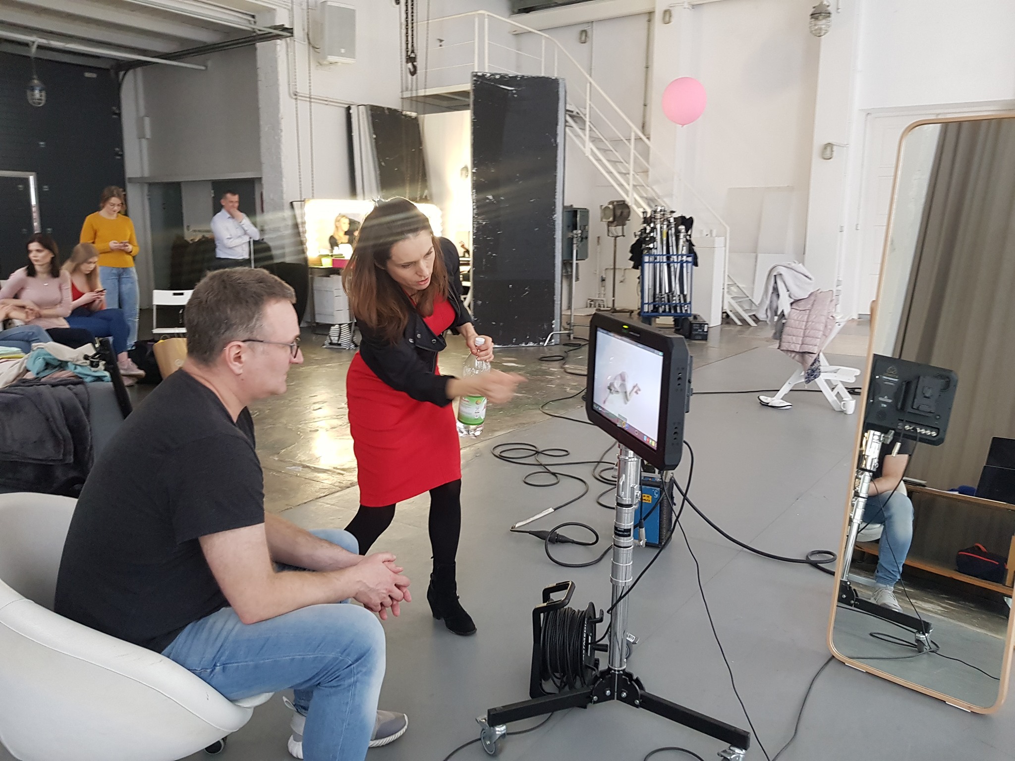 Ekipa filmowa na planie: kobieta w czerwonej sukience reżyseruje ujęcie na monitorze, aktor siedzi na krześle, w tle oświetlenie i sprzęt studyjny.