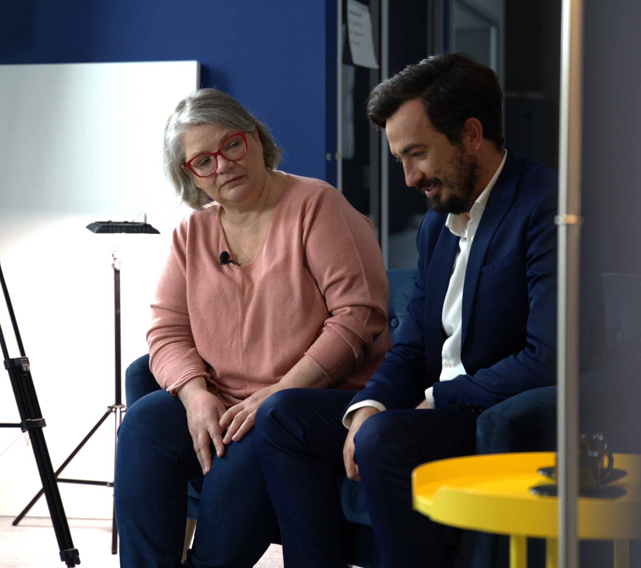 Kadr z wywiadu: kobieta w okularach z mikrofonem i mężczyzna w garniturze siedzą obok siebie, w tle sprzęt oświetleniowy i niebieska ściana.