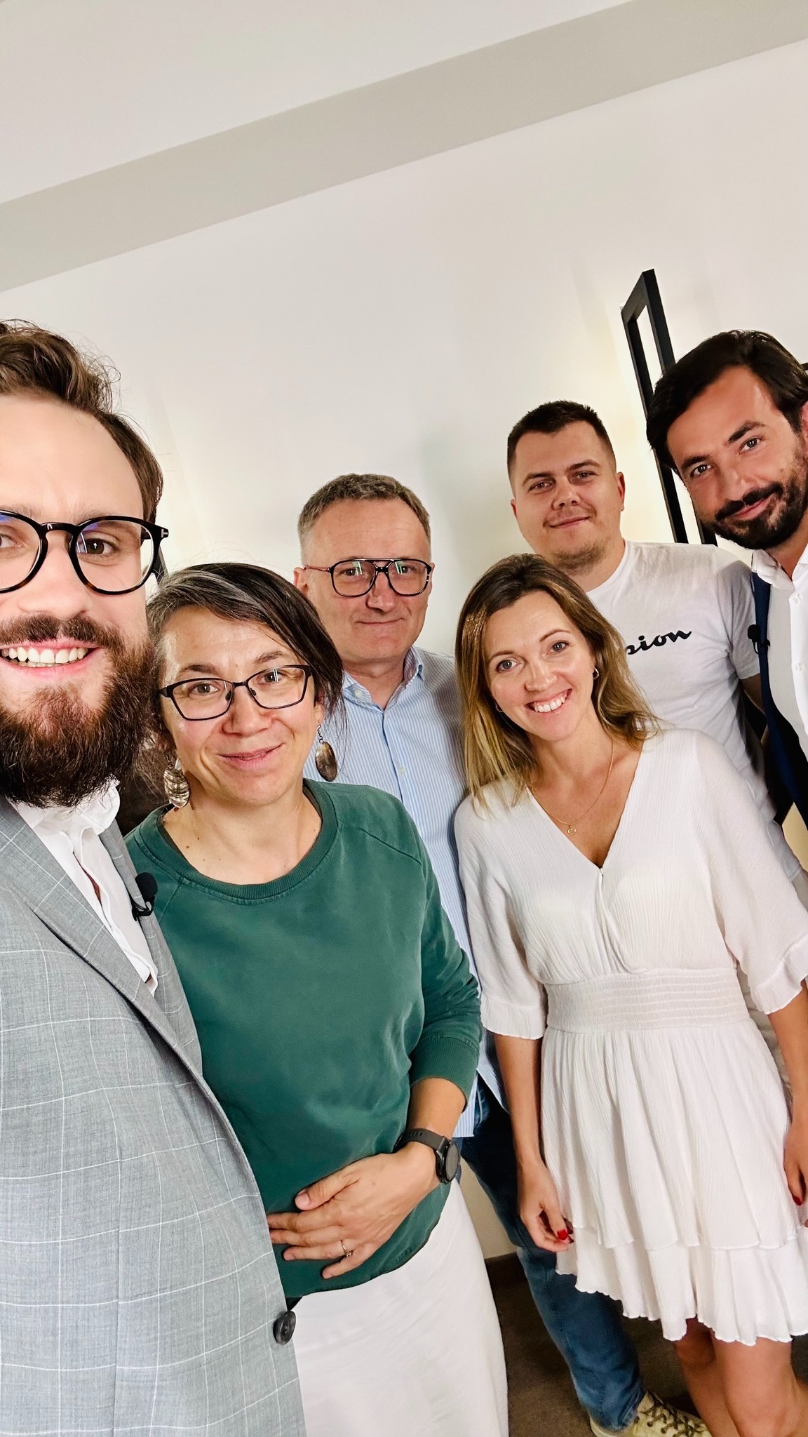 Grupowe selfie sześciu uśmiechniętych osób w różnym wieku, w jasnym pomieszczeniu z minimalistycznym wystrojem. Mężczyzna w okularach i szarej marynarce na pierwszym planie.
