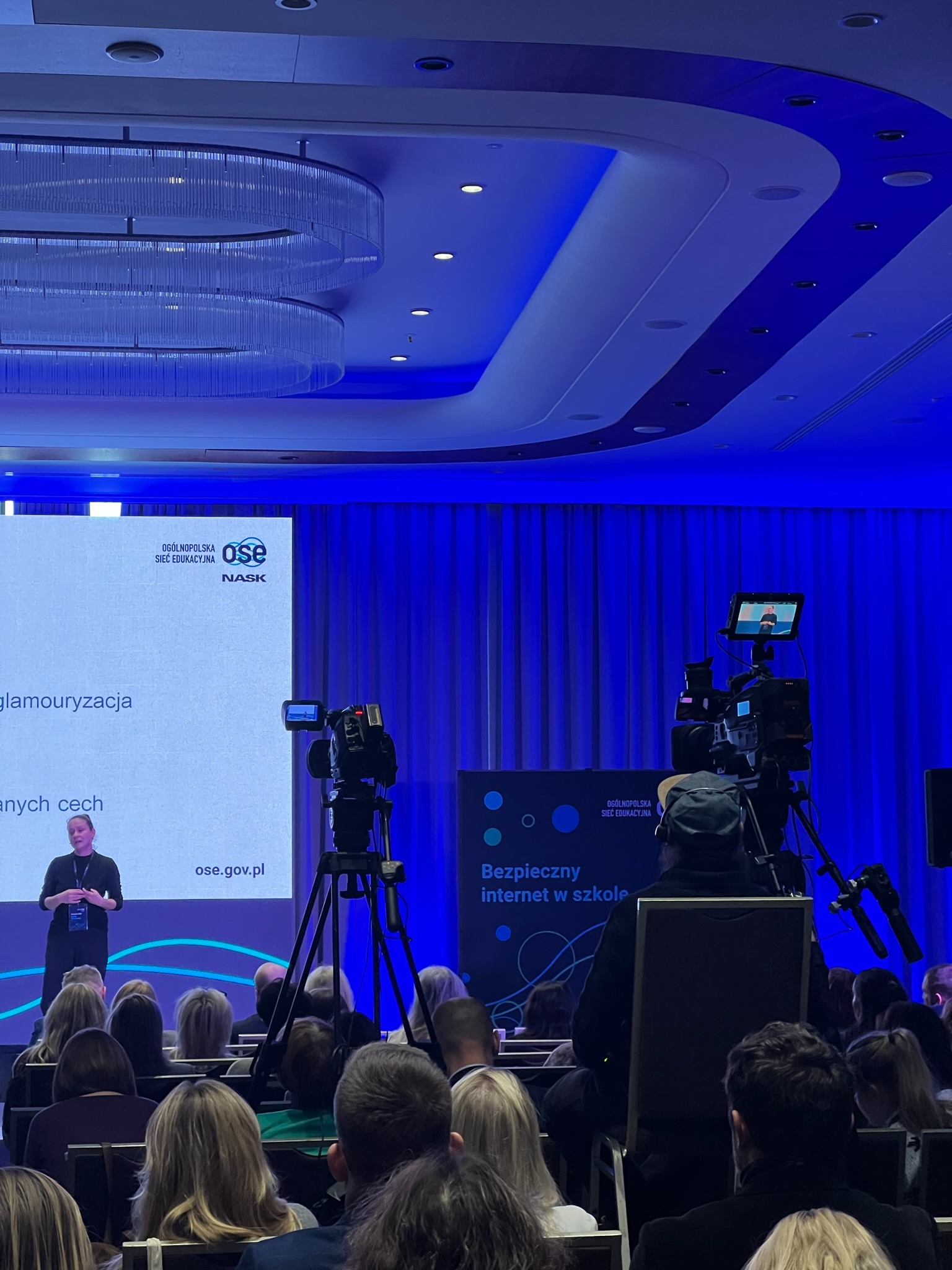 Nagranie prelekcji: kamera na statywie rejestruje prelegentkę na tle ekranu z napisem 'Bezpieczny internet w szkole' i publiczności w sali konferencyjnej.