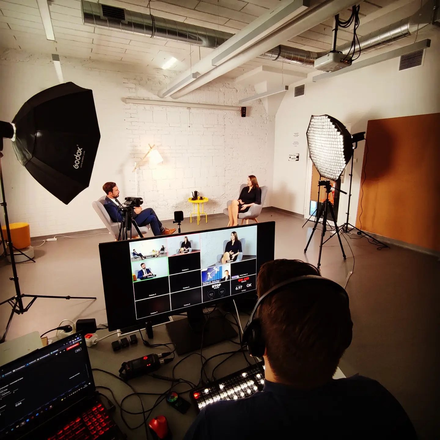 Studio nagraniowe z oświetleniem, dwoma osobami i operatorem monitorującym ujęcia na ekranie. Widoczny sprzęt: kamery, lampy, komputery.