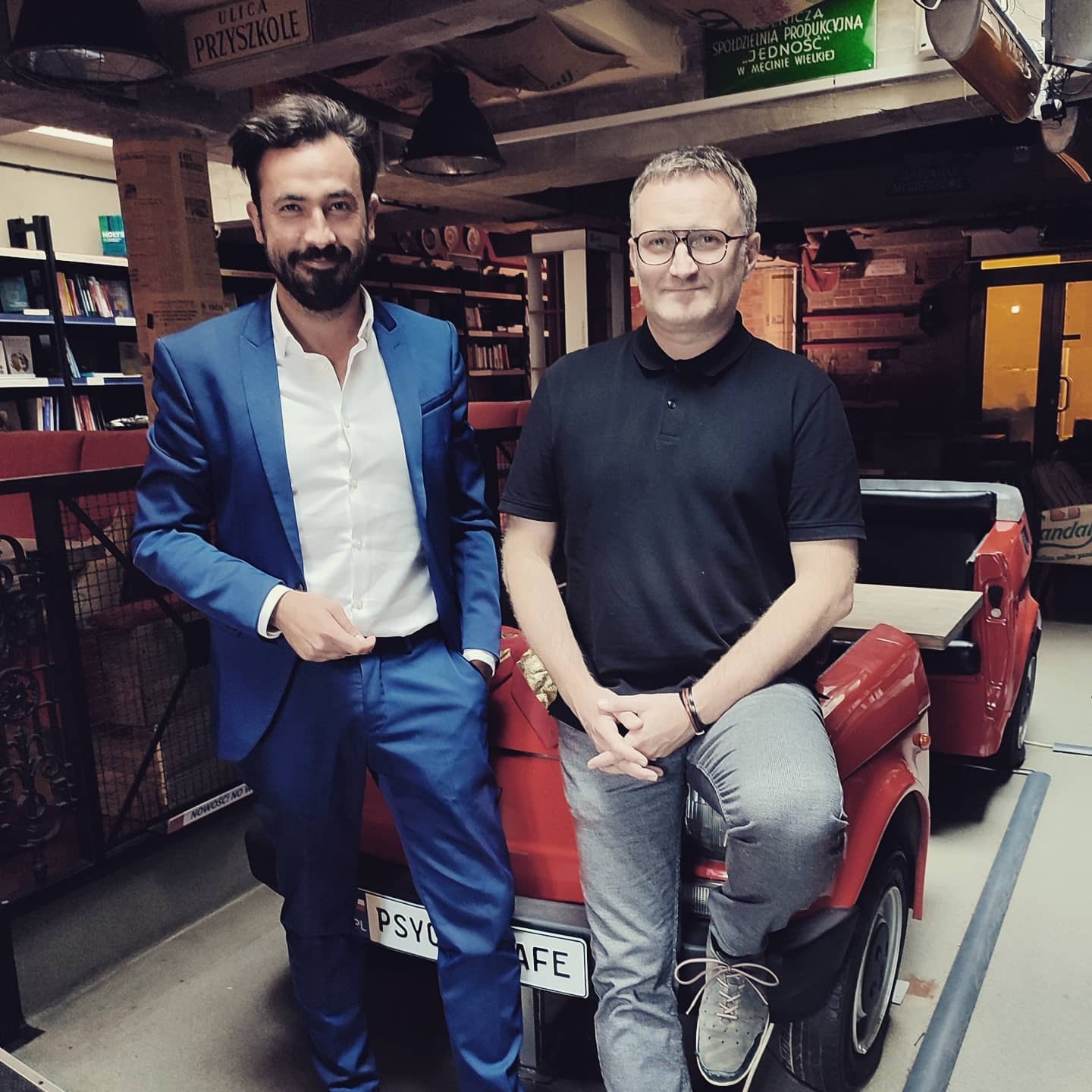 Dwóch mężczyzn pozuje na tle nietypowego wnętrza kawiarni z elementami motoryzacyjnymi, być może podczas realizacji filmu promocyjnego.