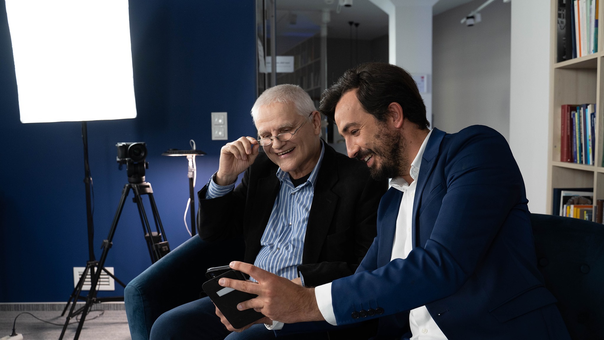 Dwóch mężczyzn w garniturach ogląda coś na smartfonie w studiu filmowym, widoczny sprzęt oświetleniowy i kamera na statywie.