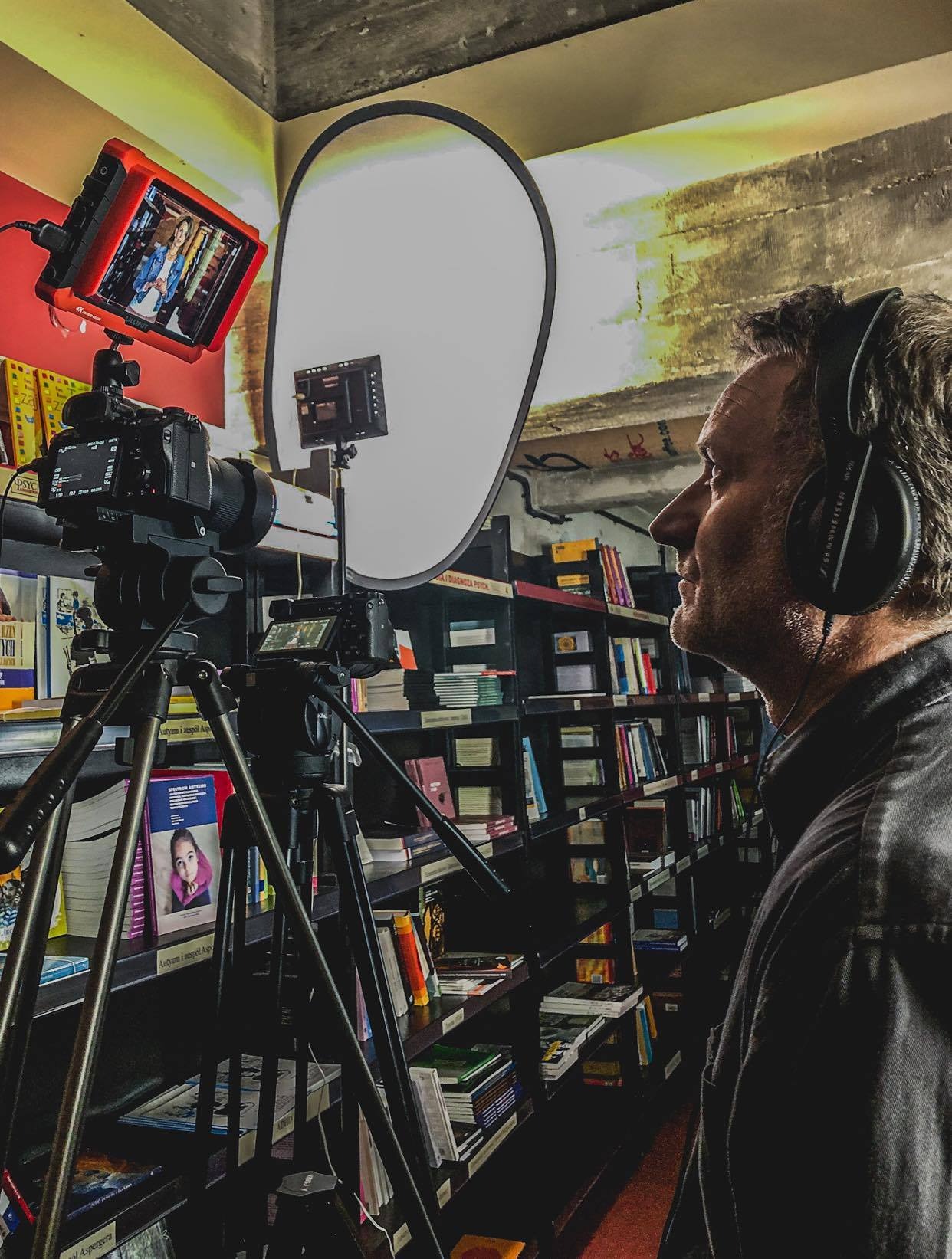Filmowiec w trakcie pracy, używa kamery na statywie i lampy oświetleniowej. W tle półki z książkami, co sugeruje nagrywanie w bibliotece lub księgarni.