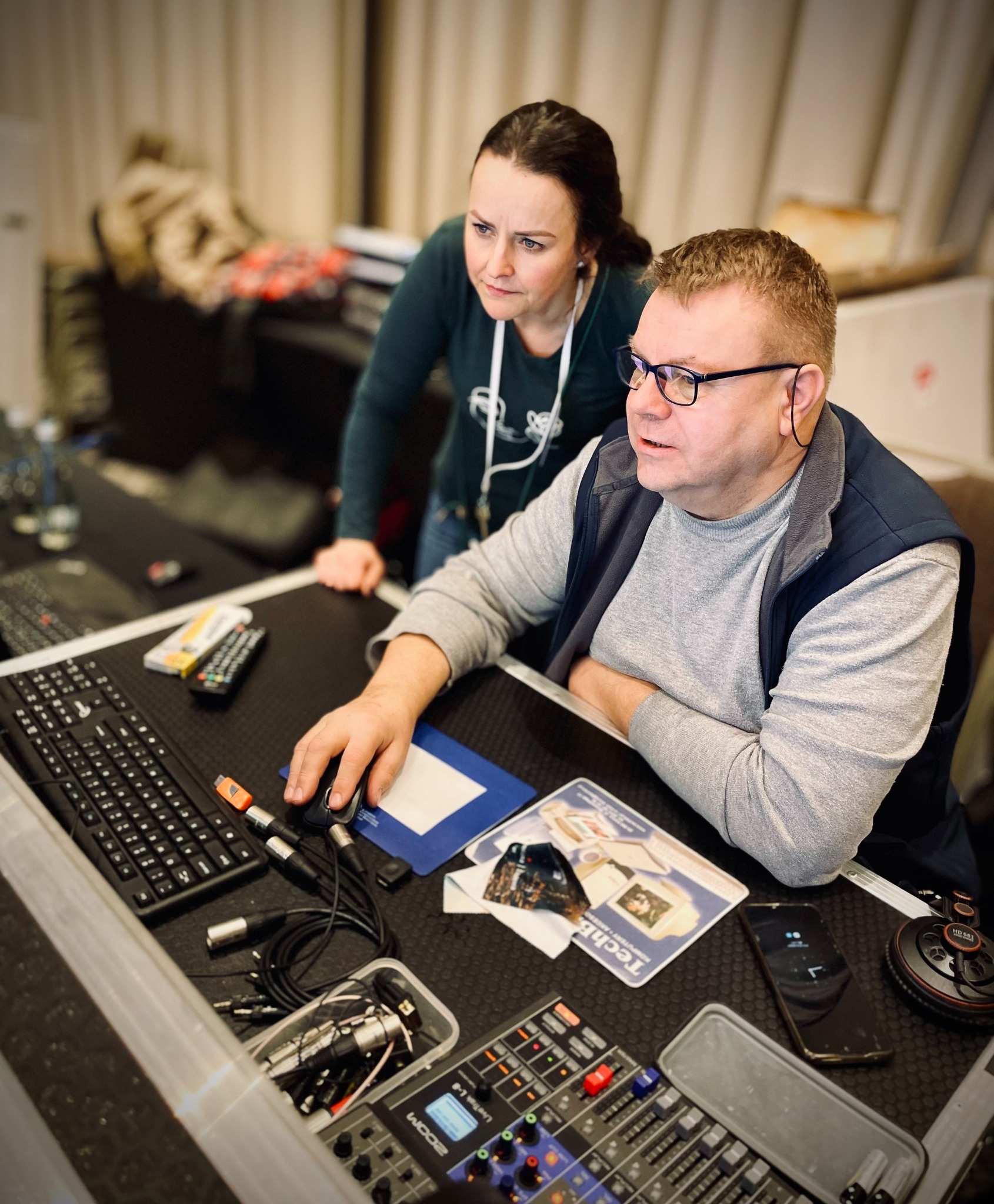 Dwóch montażystów przy stanowisku edycji wideo, mężczyzna z okularami obsługuje mikser i klawiaturę, kobieta obserwuje proces. Widoczne kable, sprzęt audio i monitor.