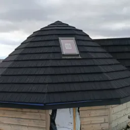 Ośmiokątna altana z czarnym dachem z gontów bitumicznych i oknem dachowym, widok na góry w tle.