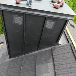 rutkos - Czarny komin wentylacyjny z kratkami na dachu pokrytym szarą dachówką bitumiczną, widoczne narzędzia i elementy montażowe, drewniana listwa wzdłuż krawędzi dachu.