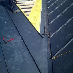 rutkos - Prace dekarskie w toku: montaż paneli na dachu z widocznymi śladami butów, żółtą membraną, narzędziami (młotek, szczypce) i fragmentem drewnianej konstrukcji.