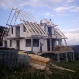 Wznoszenie dwukondygnacyjnego domu z białych bloczk&oacute;w, widoczna drewniana konstrukcja dachu w trakcie montażu, pracownik na balkonie, ułożone deski i bloczki na pierwszym planie, krajobraz w tle.