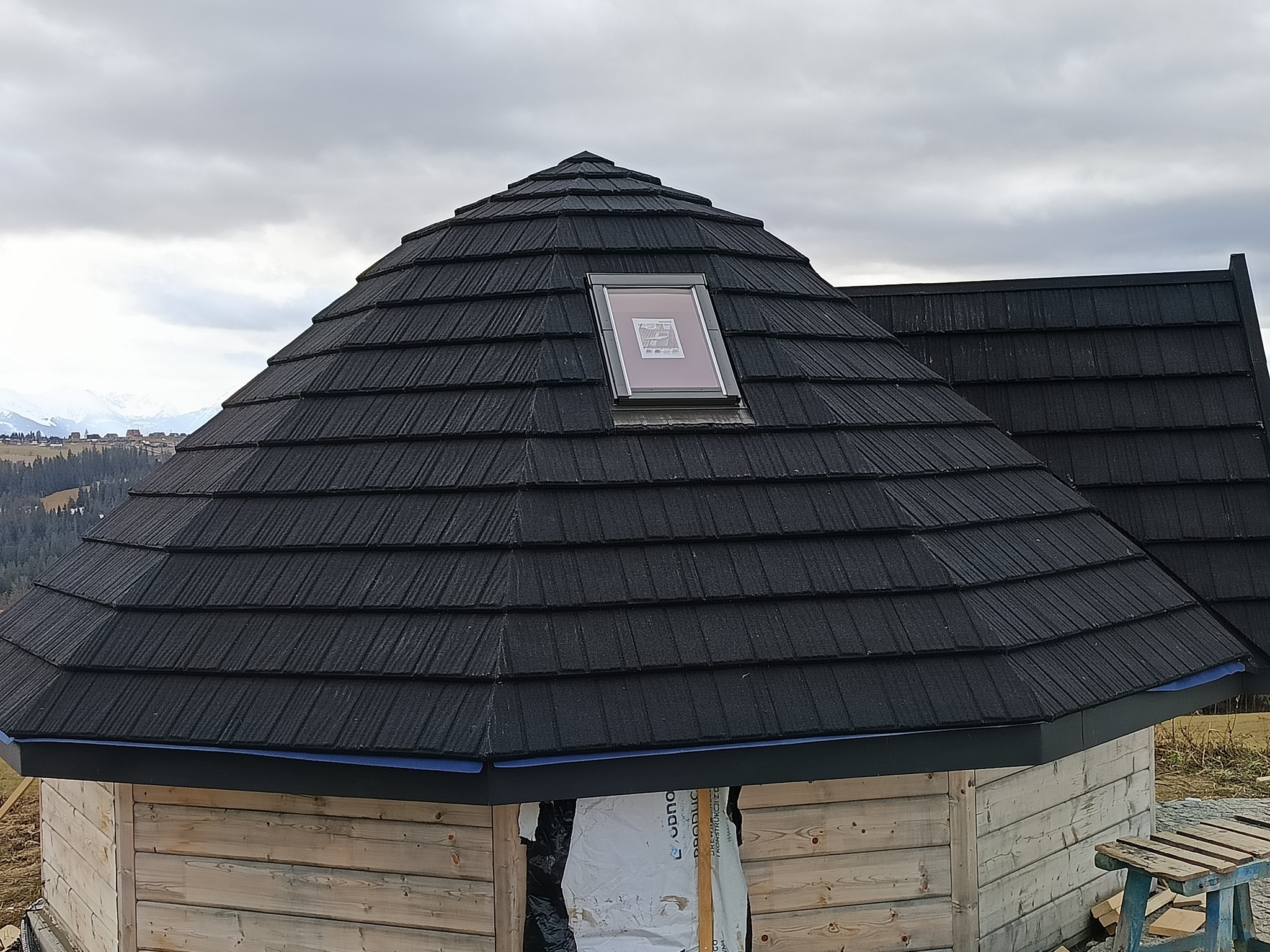 Ośmiokątna altana z czarnym dachem z gontów bitumicznych i oknem dachowym, widok na góry w tle.