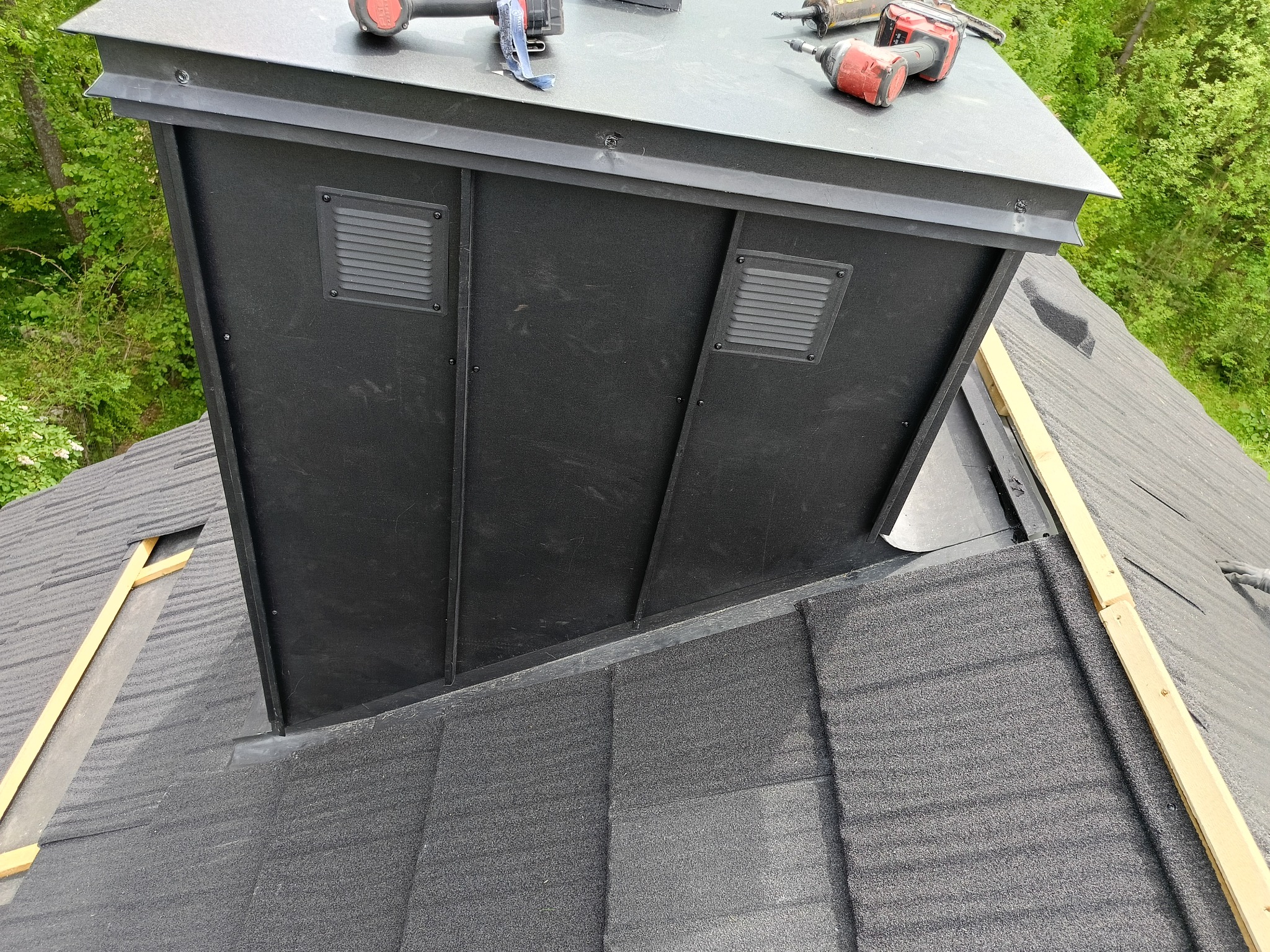 Czarny komin wentylacyjny z kratkami na dachu pokrytym szarą dachówką bitumiczną, widoczne narzędzia i elementy montażowe, drewniana listwa wzdłuż krawędzi dachu.