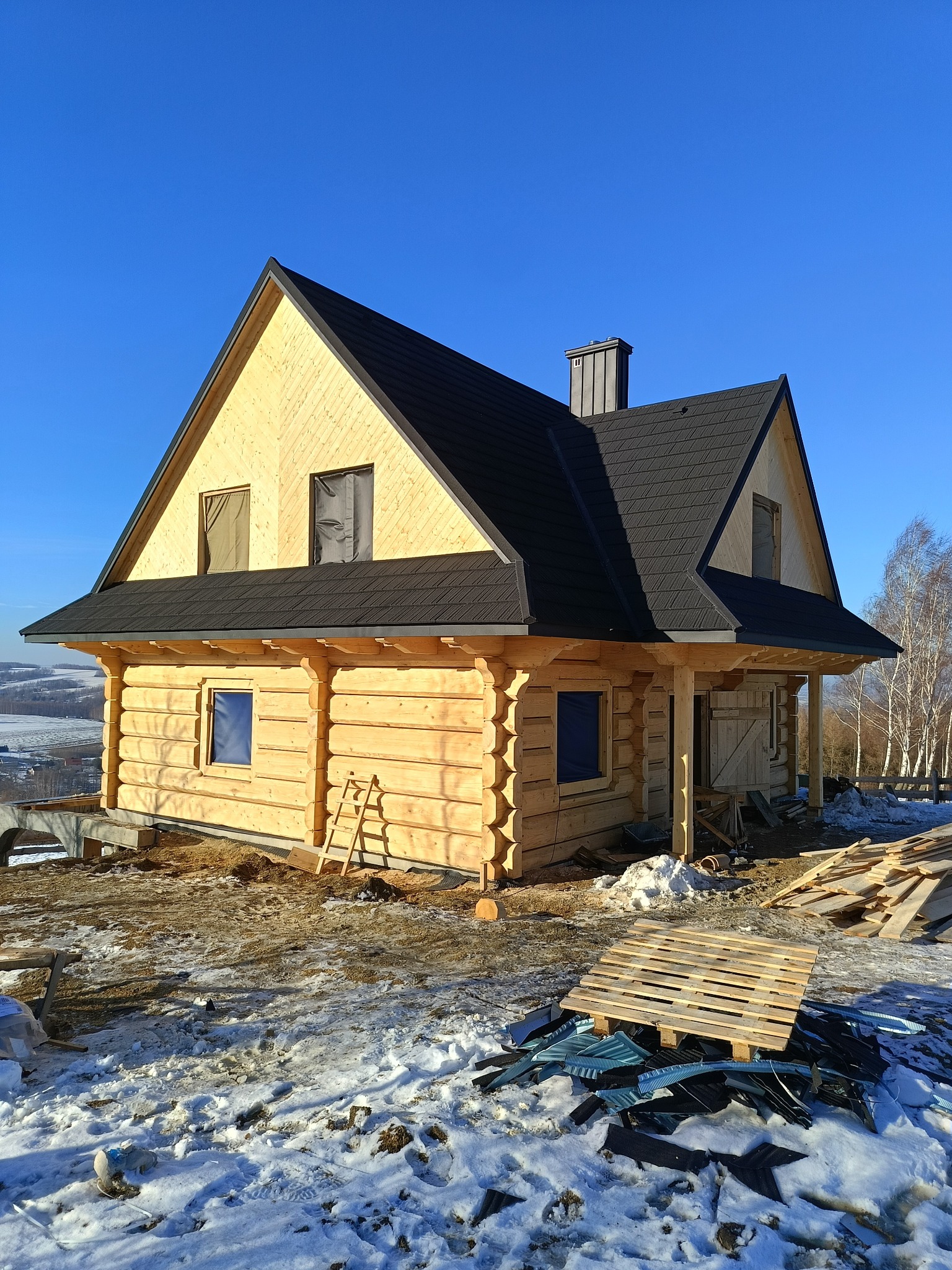 Surowy drewniany dom z ciemnym dachem z dachówki bitumicznej w trakcie budowy, widoczne zabezpieczone okna i rusztowanie, na ziemi resztki materiałów budowlanych i śnieg.