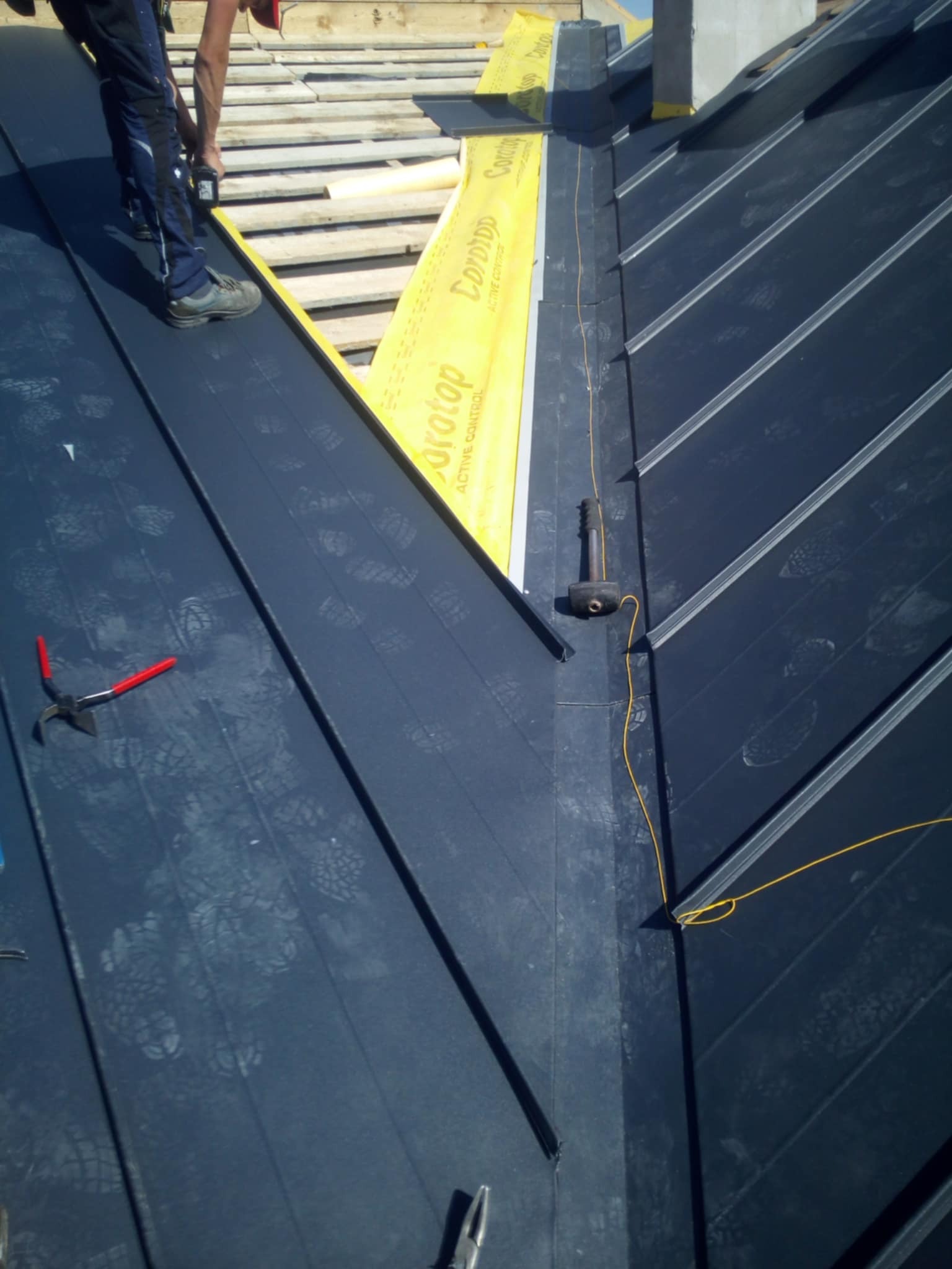 Prace dekarskie w toku: montaż paneli na dachu z widocznymi śladami butów, żółtą membraną, narzędziami (młotek, szczypce) i fragmentem drewnianej konstrukcji.