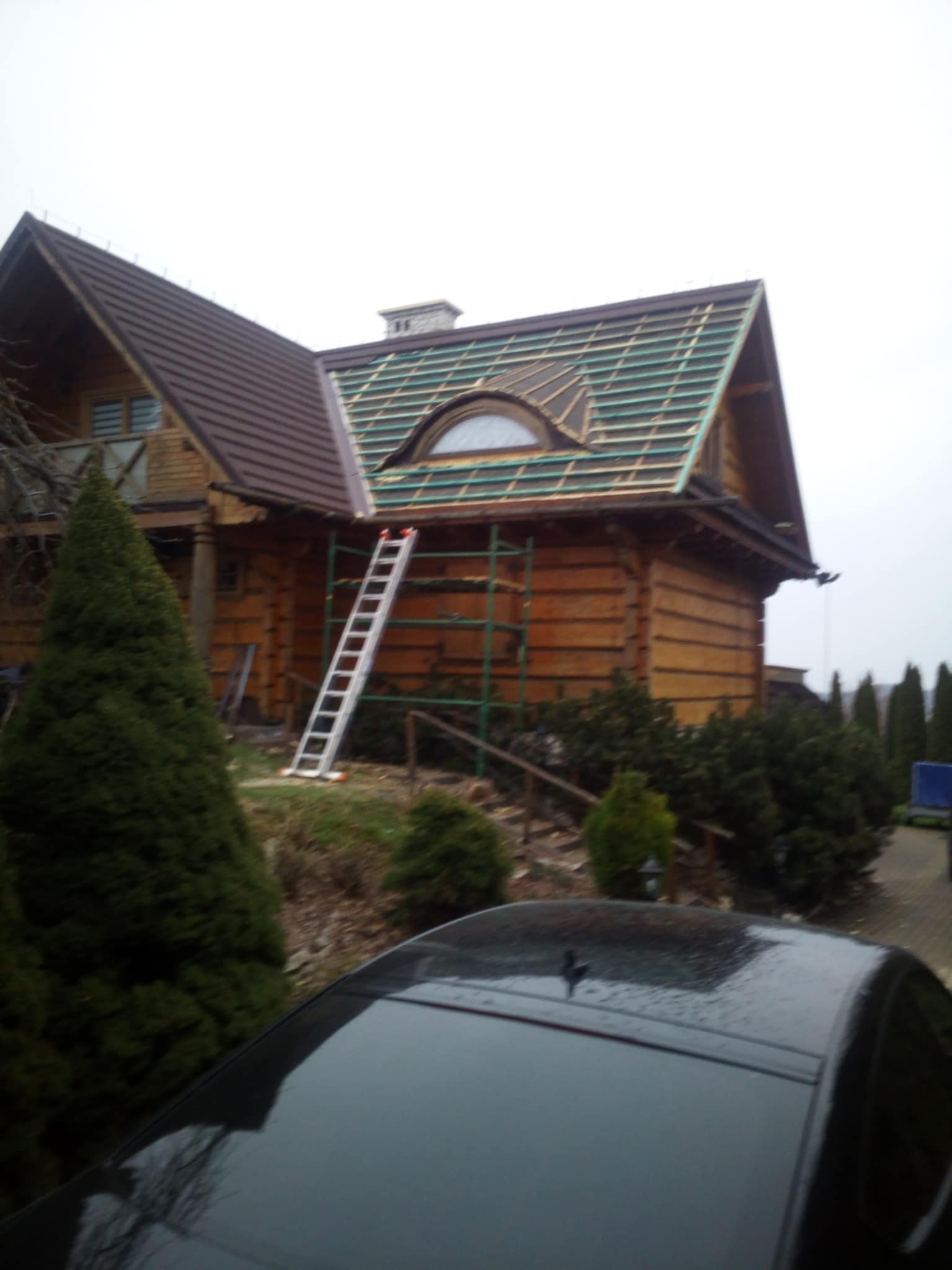 Dom z bali z częściowo wymienionym pokryciem dachowym, widoczne rusztowania i drabina oparta o ścianę, nowy dach w kolorze brązowym, stara część dachu z zielonymi łatami pod przyszłe pokrycie.