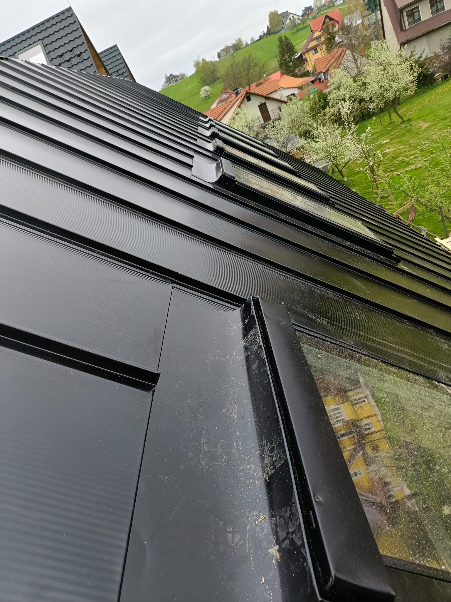 Nowy, czarny dach z blachy trapezowej z oknem dachowym, widok z bliska, z widocznymi elementami mocującymi i odbiciem otoczenia w oknie dachowym.