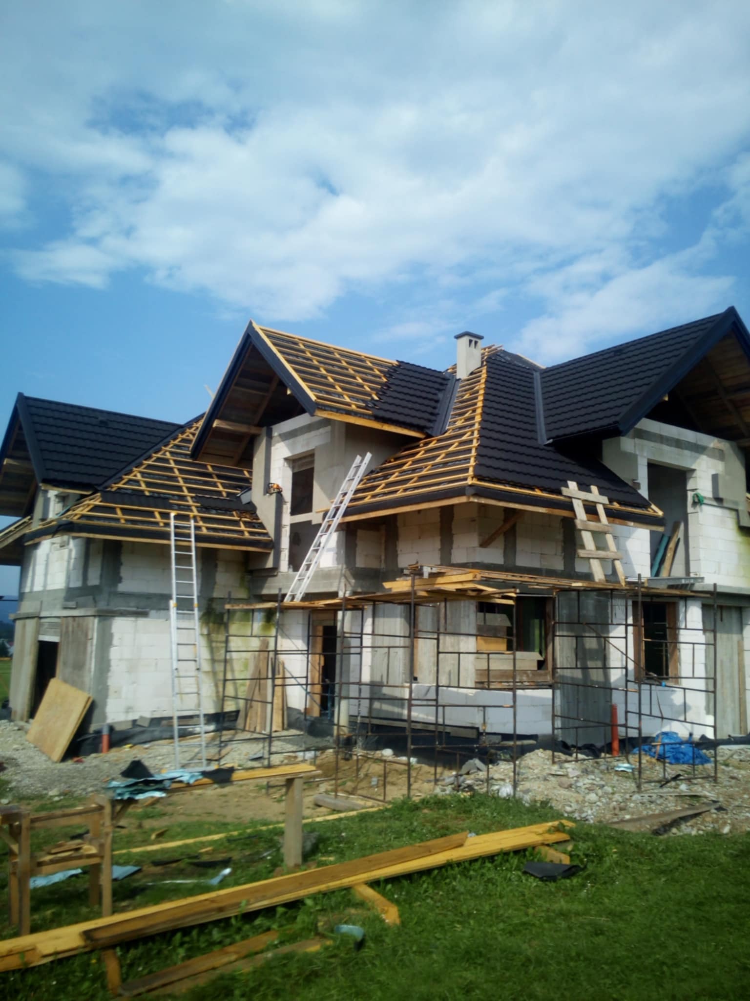 Dom w trakcie budowy z częściowo ukończonym czarnym pokryciem dachowym, widoczna więźba dachowa, rusztowania i drabiny, w tle błękitne niebo z chmurami.