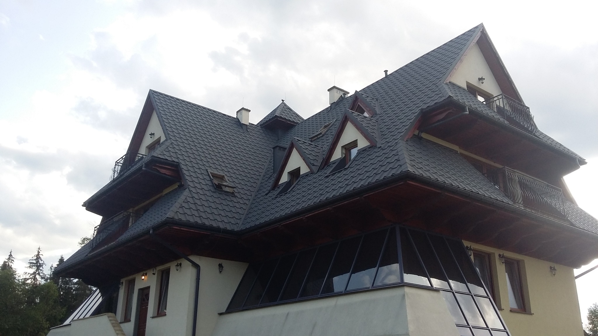 Wielopoziomowy dom z ciemnoszarą, metalową dachówką, małymi balkonami z kutymi balustradami i przeszklonym ogrodem zimowym u podstawy.