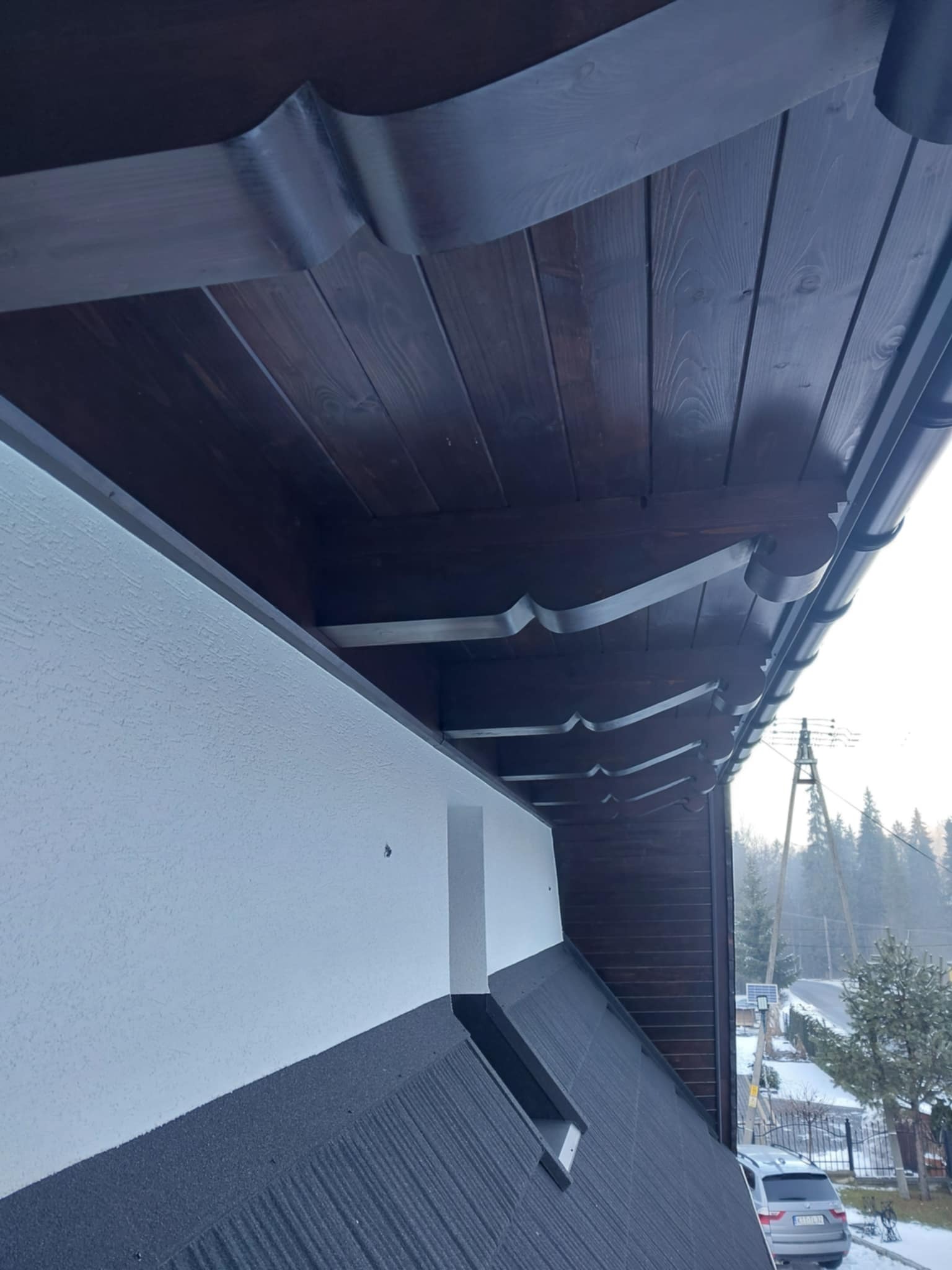 Fragment dachu z widoczną drewnianą podbitką z ozdobnymi wykończeniami, rynną i fragmentem elewacji w zimowej scenerii.