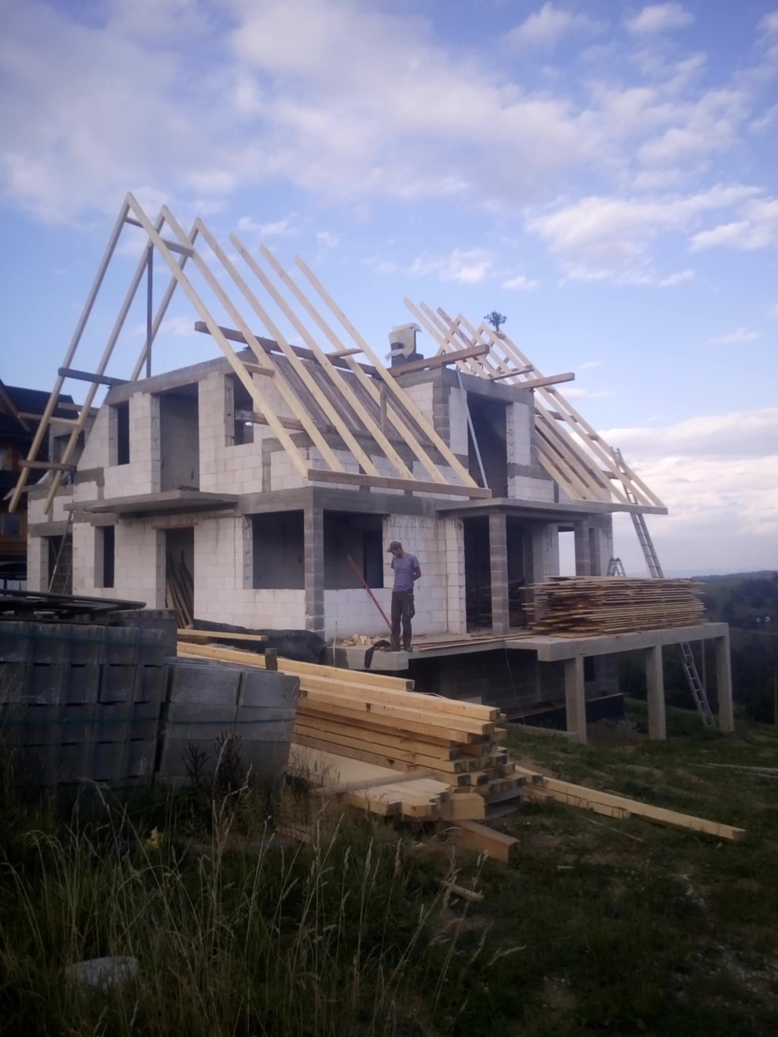 Wznoszenie dwukondygnacyjnego domu z białych bloczków, widoczna drewniana konstrukcja dachu w trakcie montażu, pracownik na balkonie, ułożone deski i bloczki na pierwszym planie, krajobraz w tle.